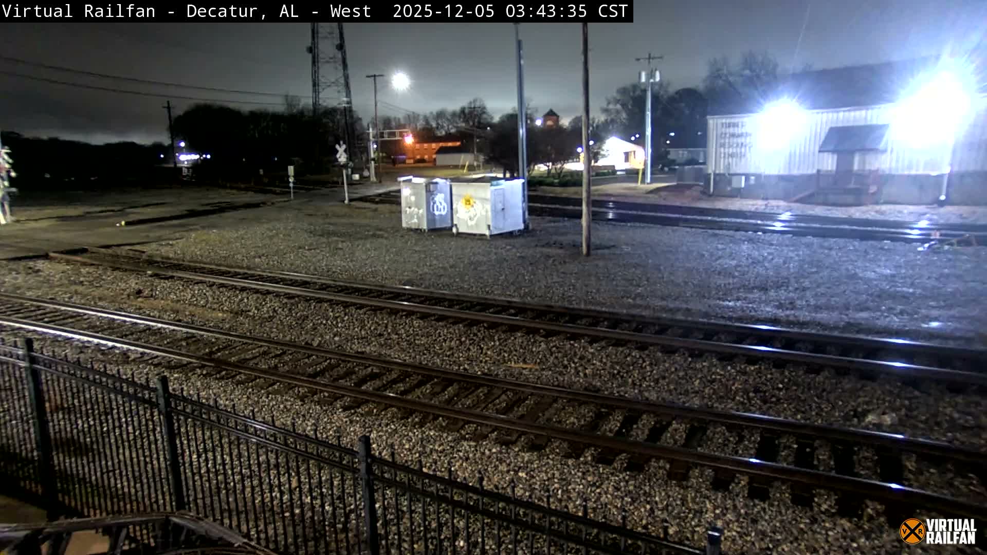 Multiple train tracks run through an artificially lit rail yard at night under a clear sky, with a black metal fence in the foreground and industrial buildings, utility poles, and streetlights in the background.