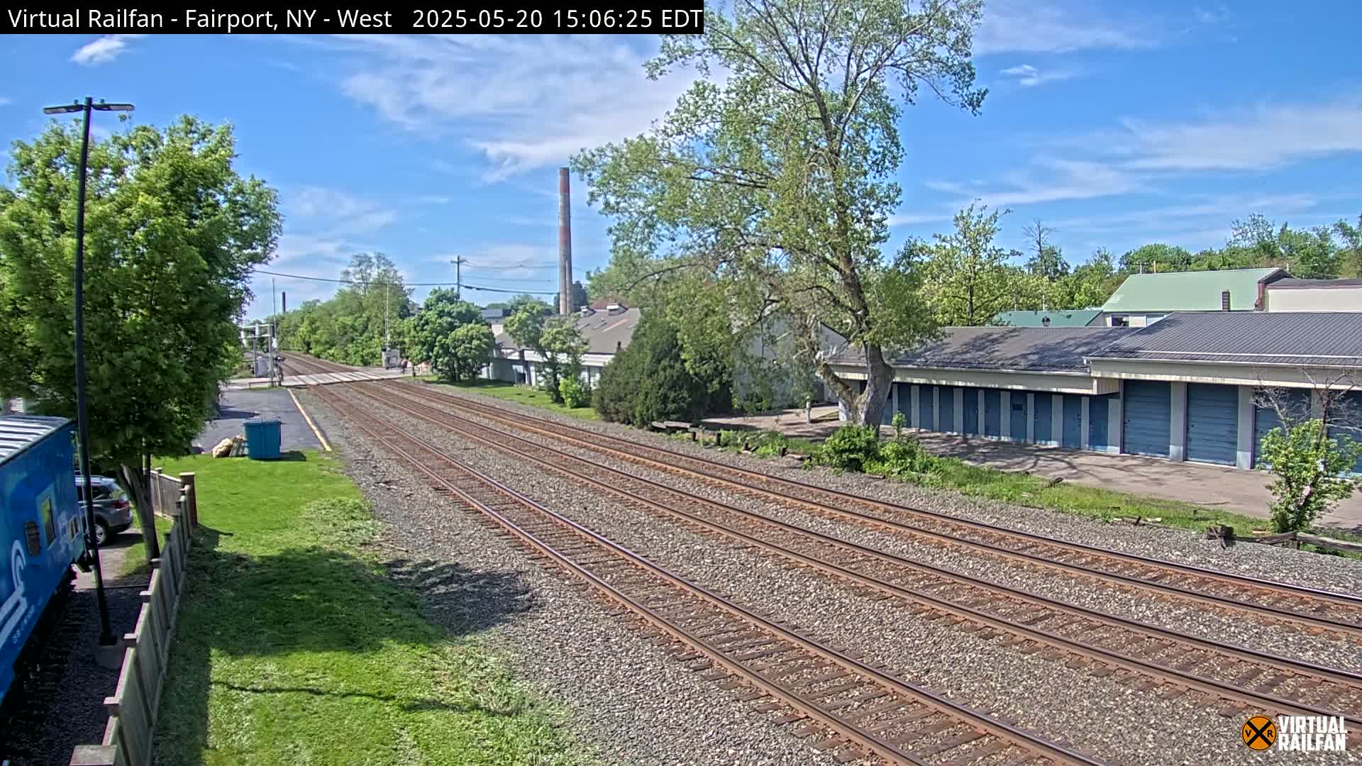 Multiple train tracks stretch into the distance under a partly sunny sky, passing alongside buildings and lush green vegetation.