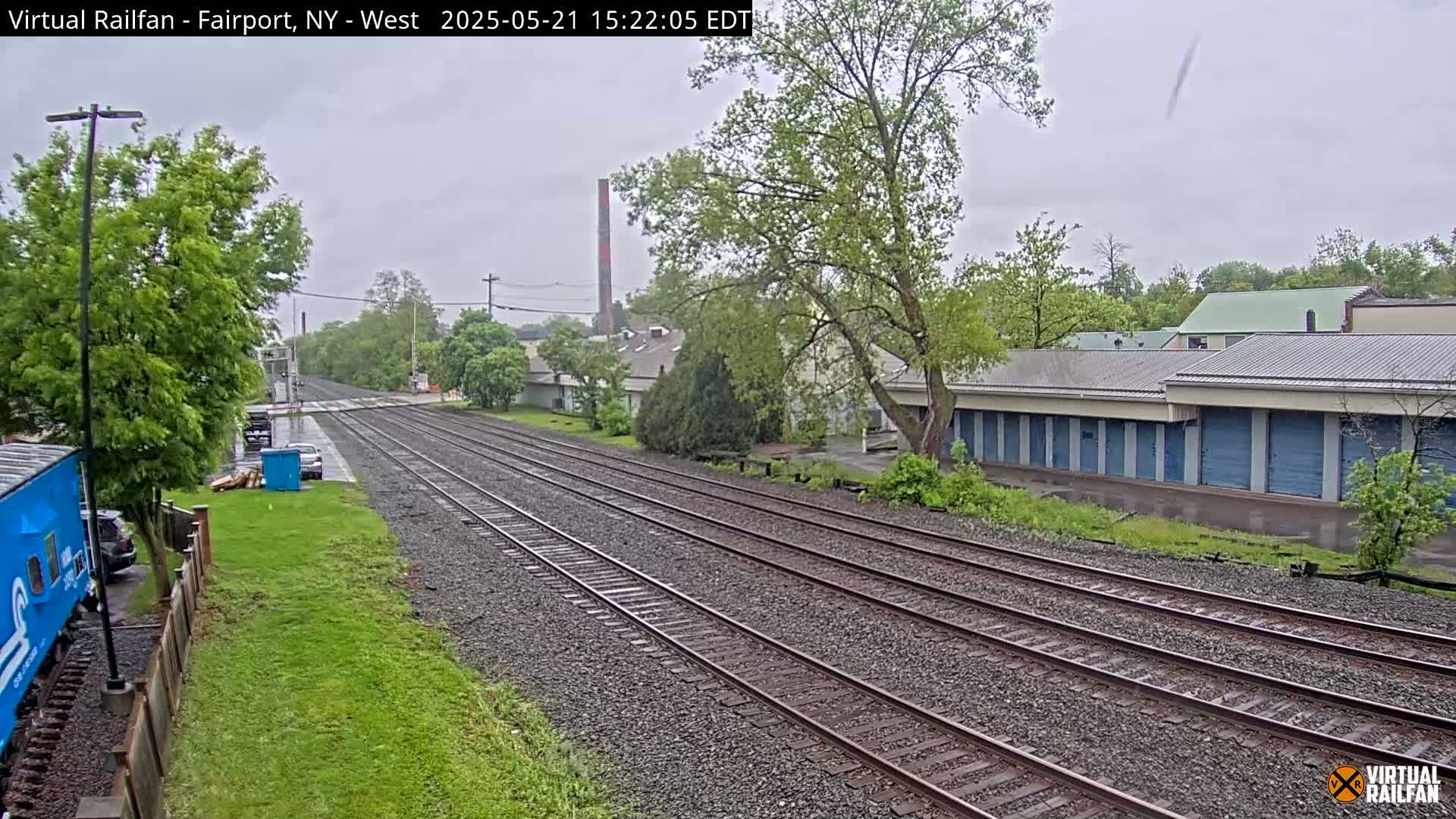 Multiple train tracks stretch into the distance on an overcast day, flanked by greenery, buildings, and a few parked vehicles.