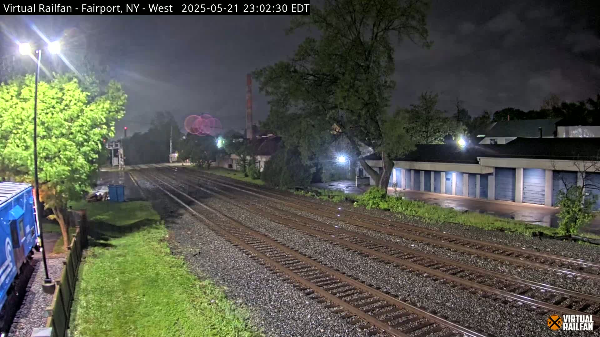 A nighttime view of rain-slicked railroad tracks extending into the distance, past a row of storage units and under a partly cloudy sky.