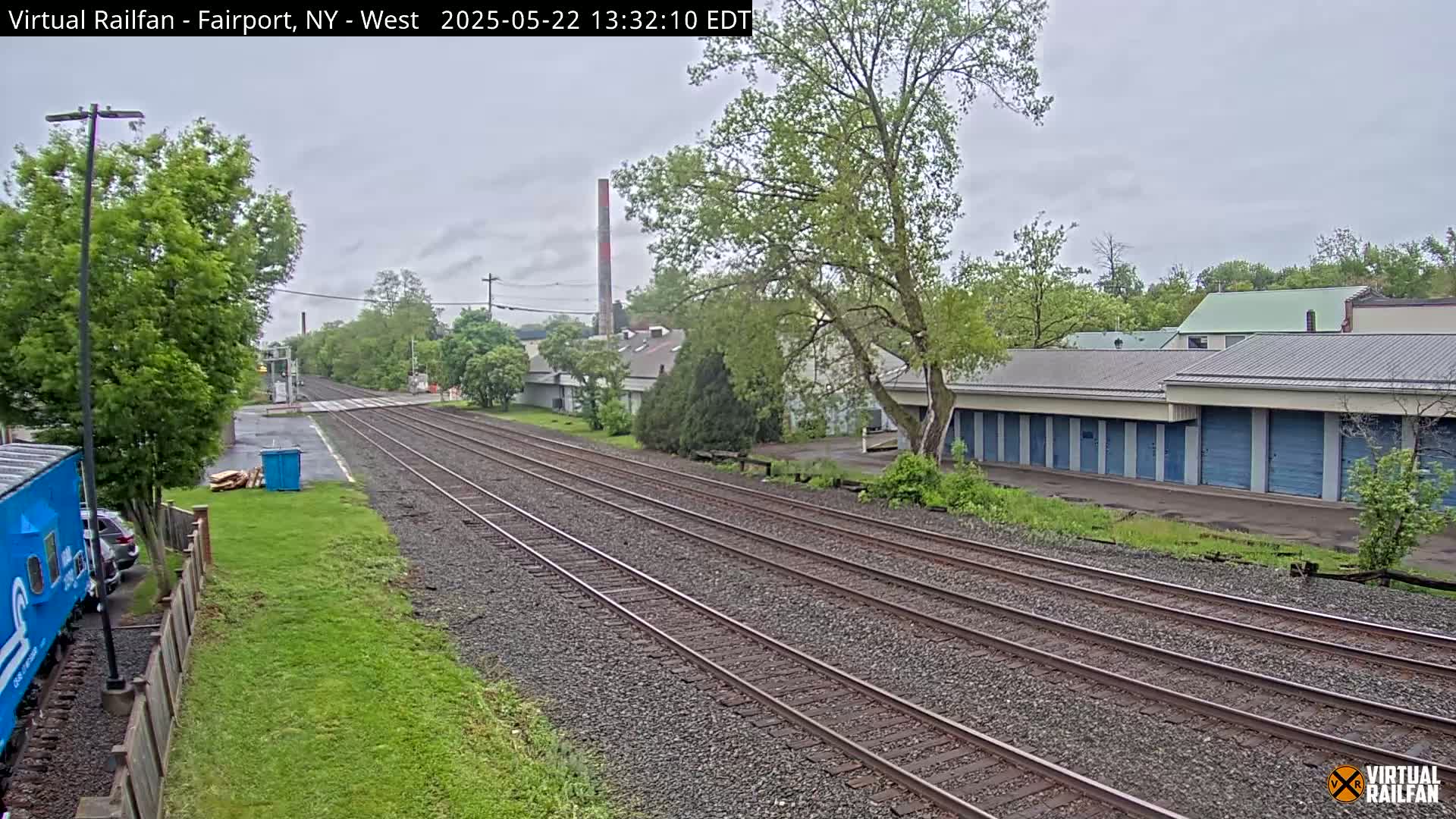 A view of railroad tracks on an overcast day, extending from the foreground to a row of storage units and industrial buildings beyond, with lush green vegetation lining the sides.