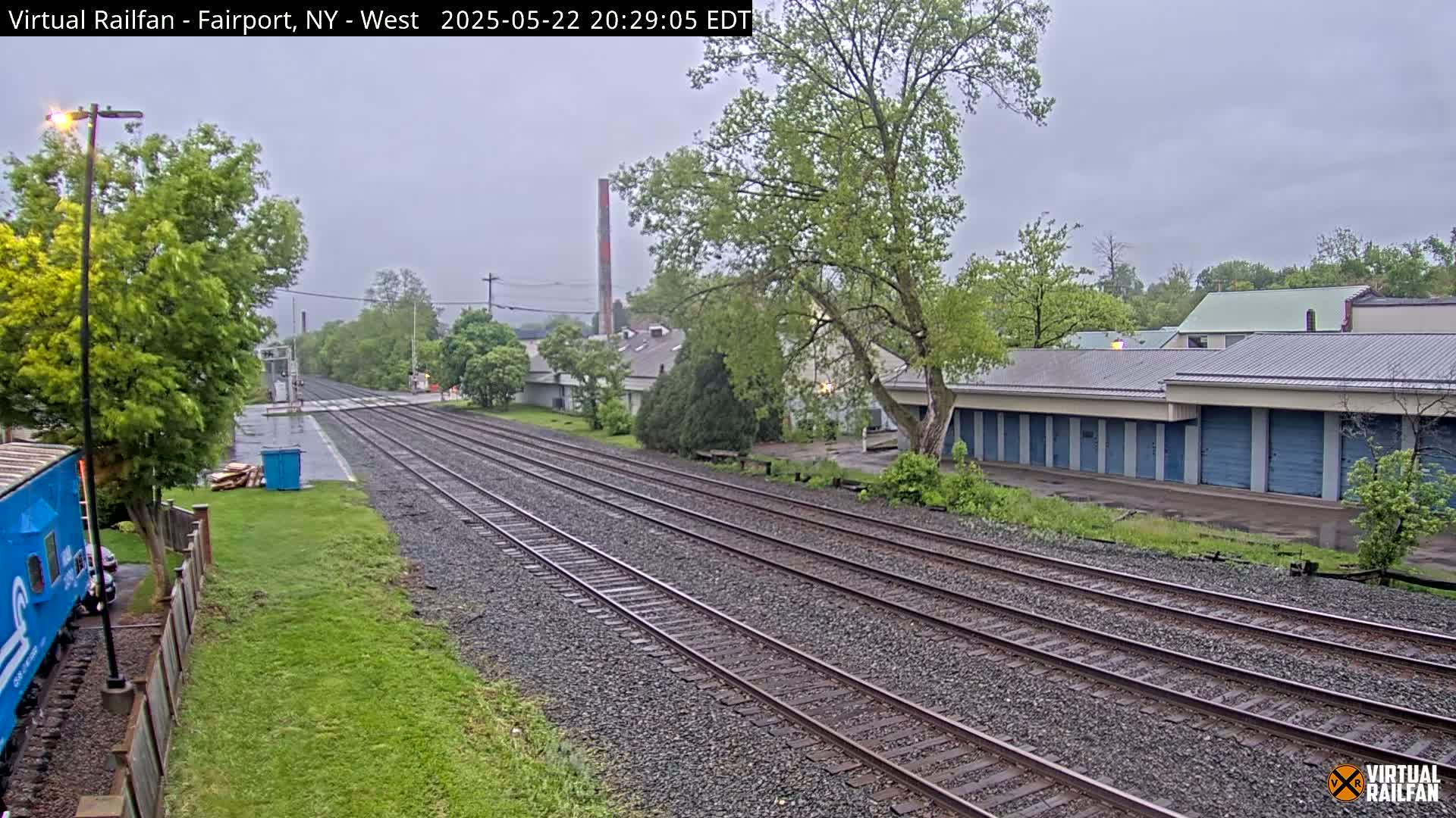 A view of multiple train tracks on a overcast day, with a row of storage units and some greenery to the right, and a partial view of a blue train car to the left.