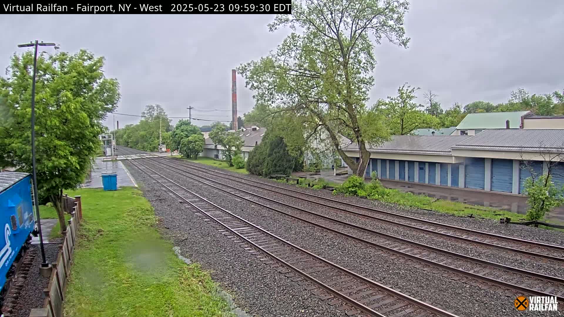 Multiple sets of train tracks stretch into the distance on an overcast day, past a row of storage units and other buildings, under a grey sky.