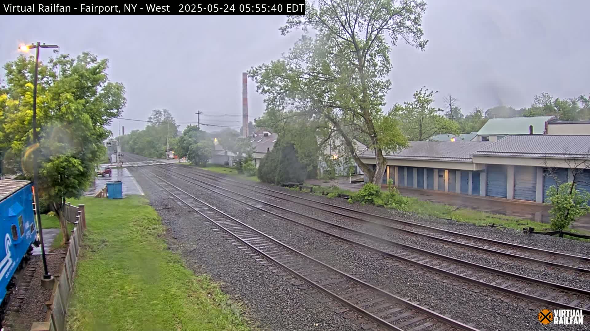 A view of railroad tracks on a rainy day, with trees, buildings, and a storage facility visible alongside.