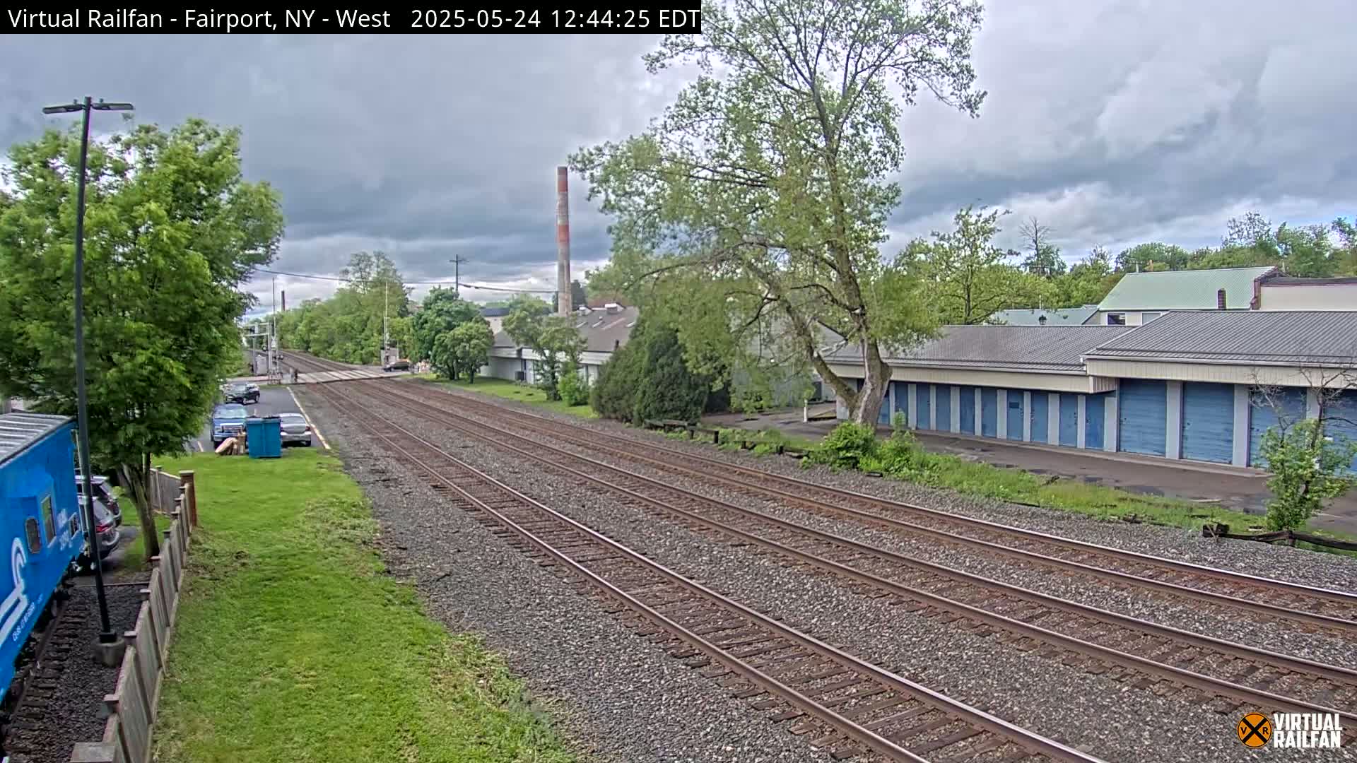 A view of train tracks under a cloudy sky, alongside a row of blue storage units and some parked cars near leafy green trees and buildings.
