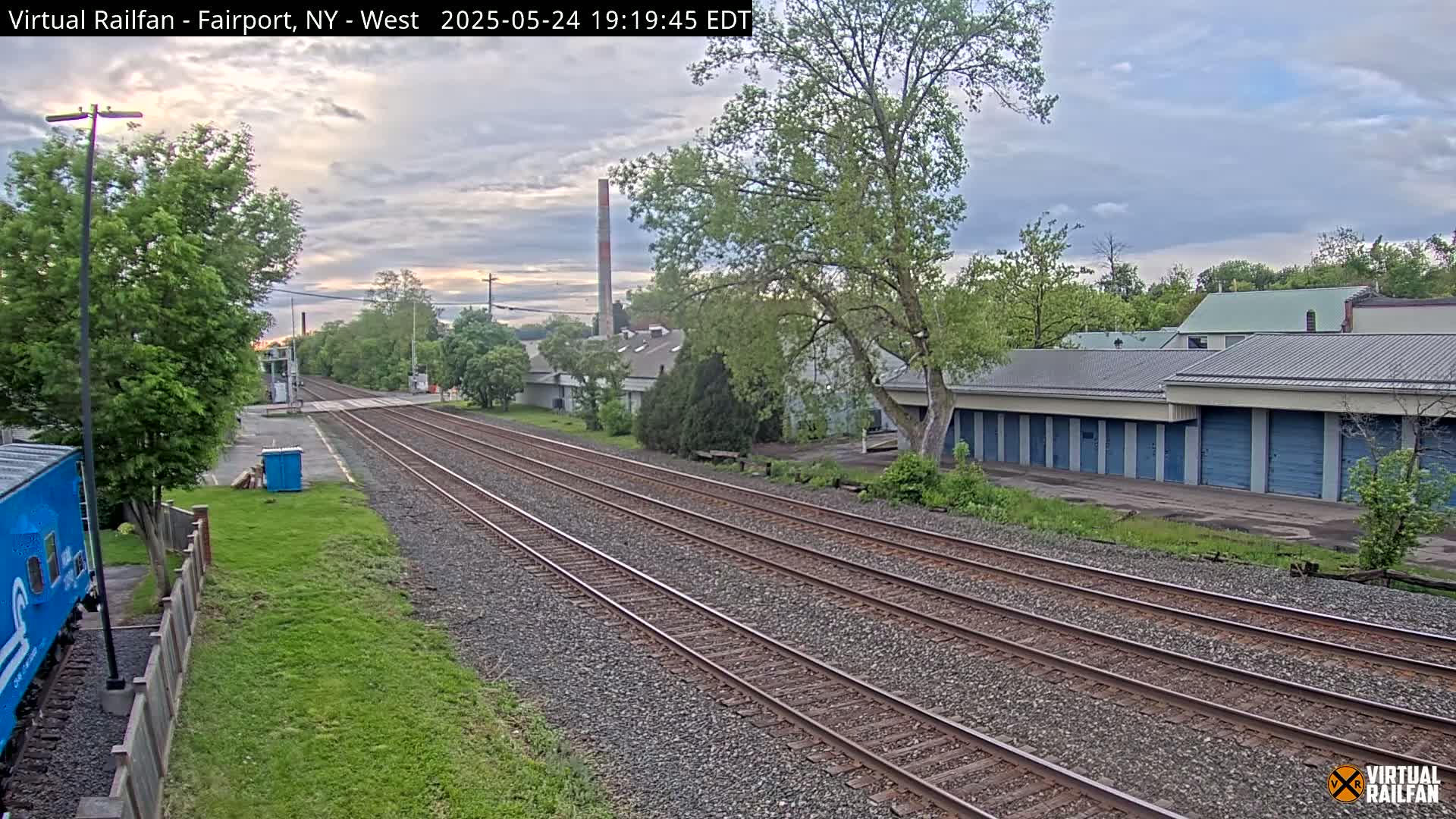 A view of train tracks extending into the distance under a partly cloudy sky, with buildings and greenery lining the sides.