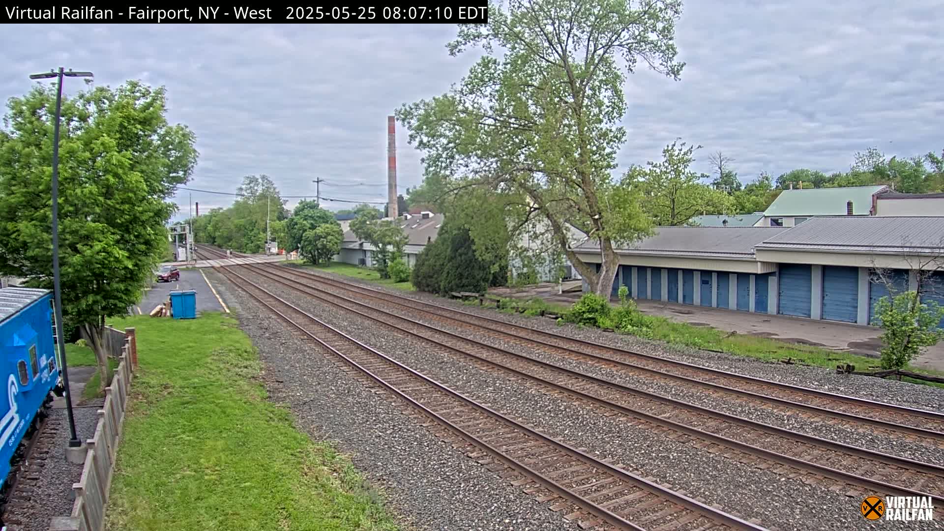 Three parallel train tracks extend into the distance past a row of storage units and some leafy green trees under an overcast sky.