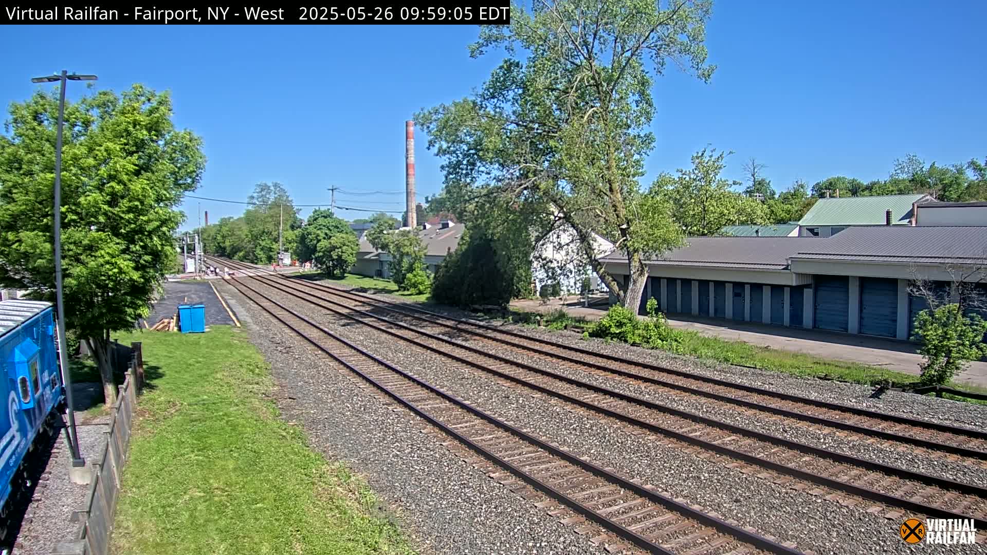 Multiple train tracks extend into the distance on a sunny day, passing alongside industrial buildings and lush green vegetation.