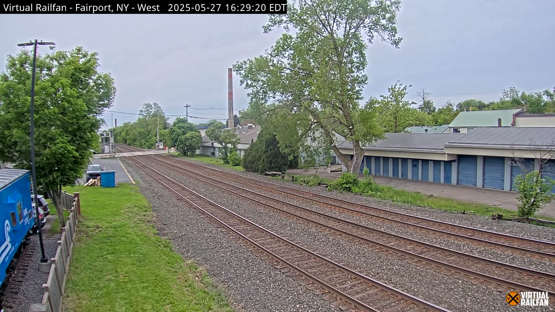 A view of railroad tracks extending into the distance on an overcast day, passing alongside a row of storage units and other buildings, with lush green vegetation surrounding the scene.