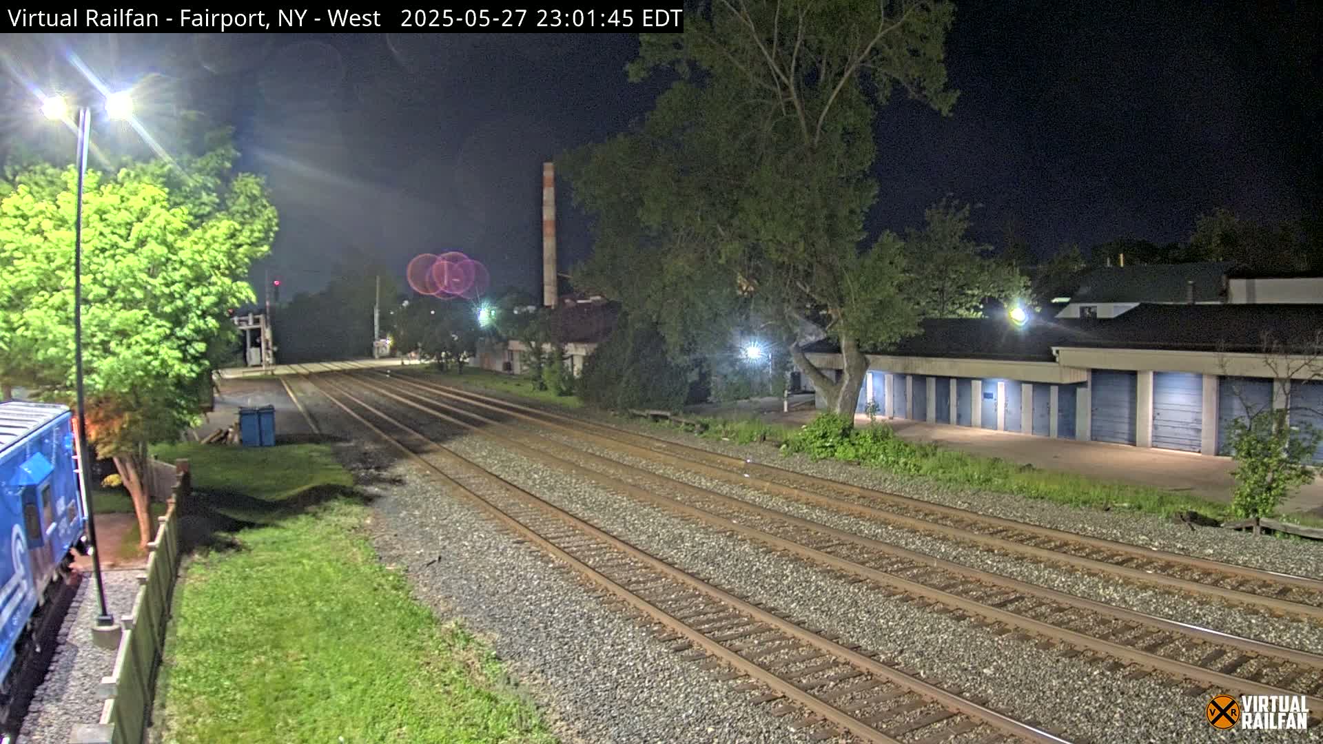 At night, a train is partially visible near multiple sets of train tracks that extend toward a row of storage units under a dark sky.