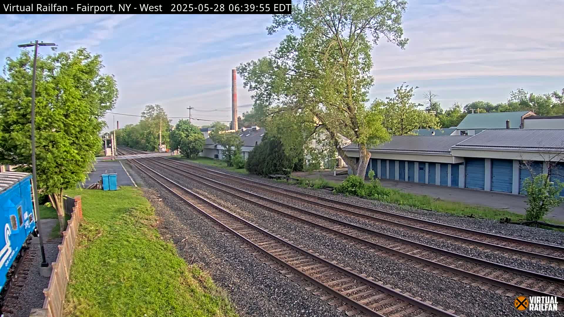 Three sets of train tracks stretch into the distance under a partly cloudy sky, passing alongside a row of storage units and industrial buildings.