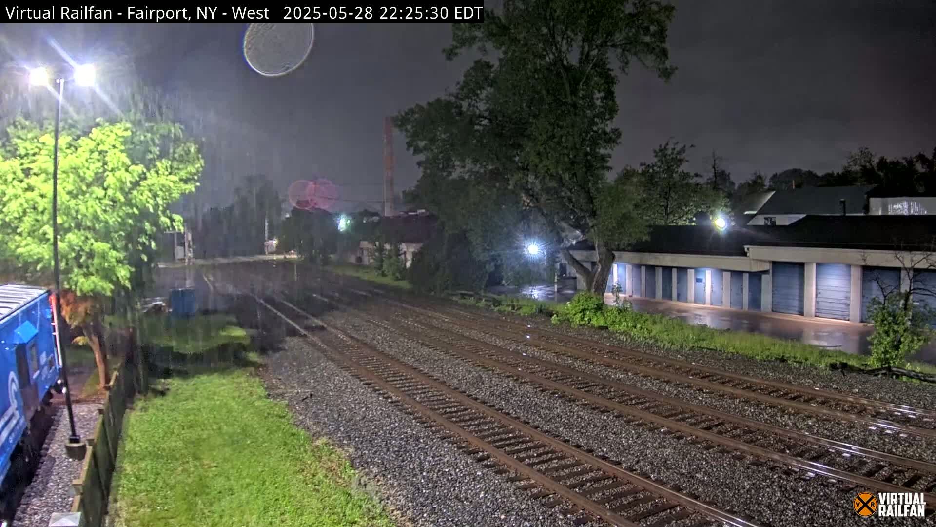 A nighttime view of railroad tracks during heavy rain, with a row of storage units and lush greenery visible on either side.