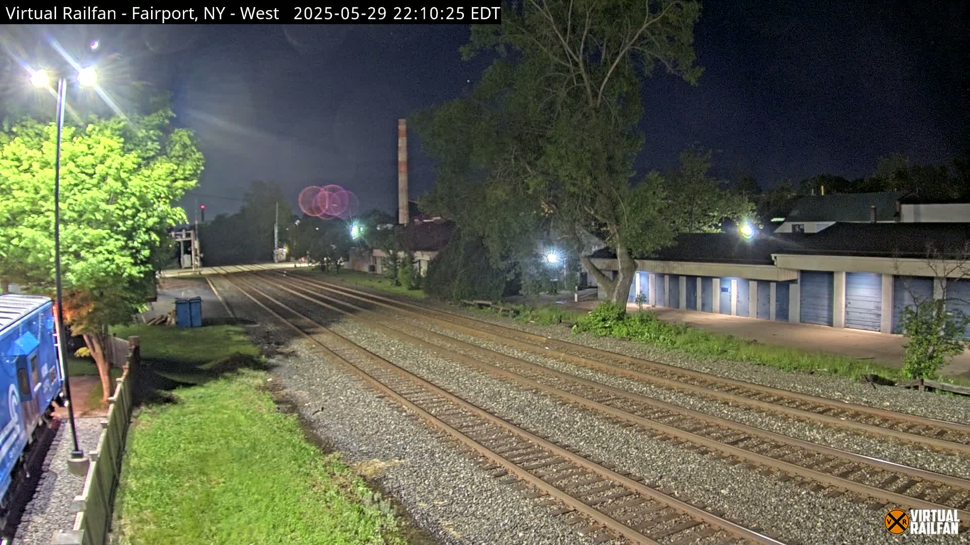 A nighttime view of train tracks extending into the distance, flanked by grassy areas, a row of storage units, and trees under a clear, dark sky.