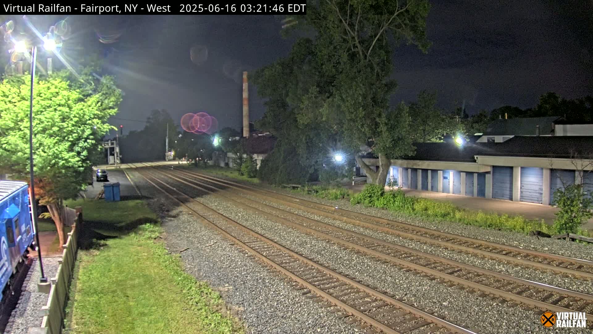 Multiple sets of train tracks at night, next to a row of storage units and some trees under a dark sky.