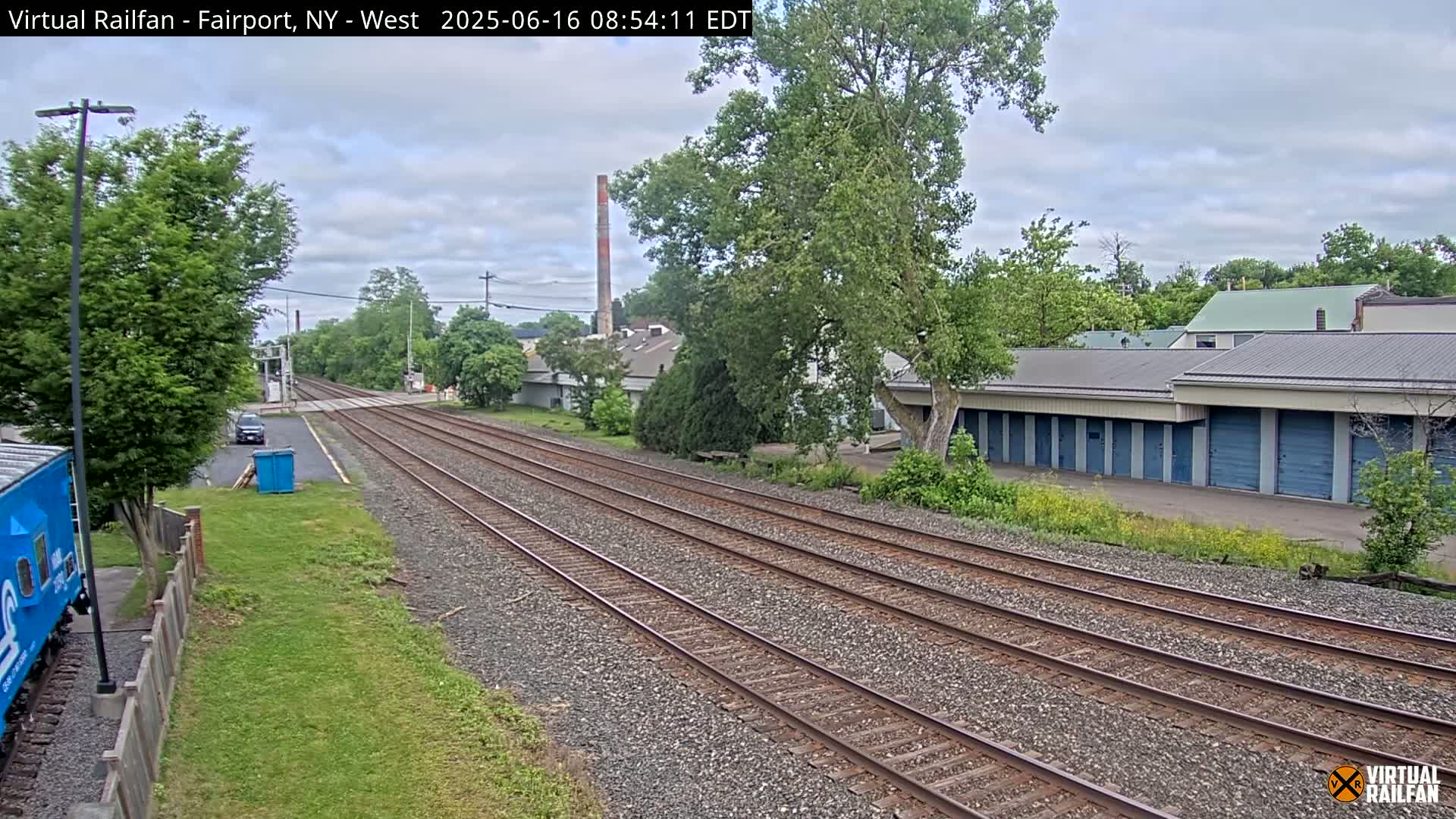Multiple train tracks stretch into the distance, passing beside a row of storage units and some trees under a partly cloudy sky.
