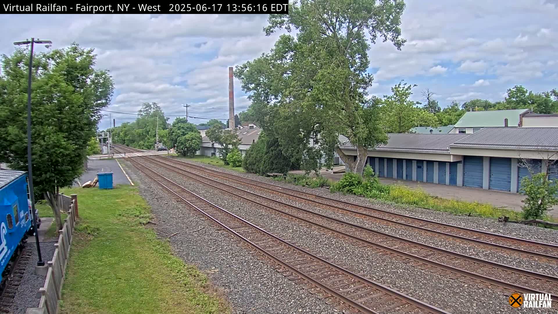 Multiple train tracks stretch into the distance under a partly cloudy sky, passing alongside a row of storage units and other buildings near lush green vegetation.