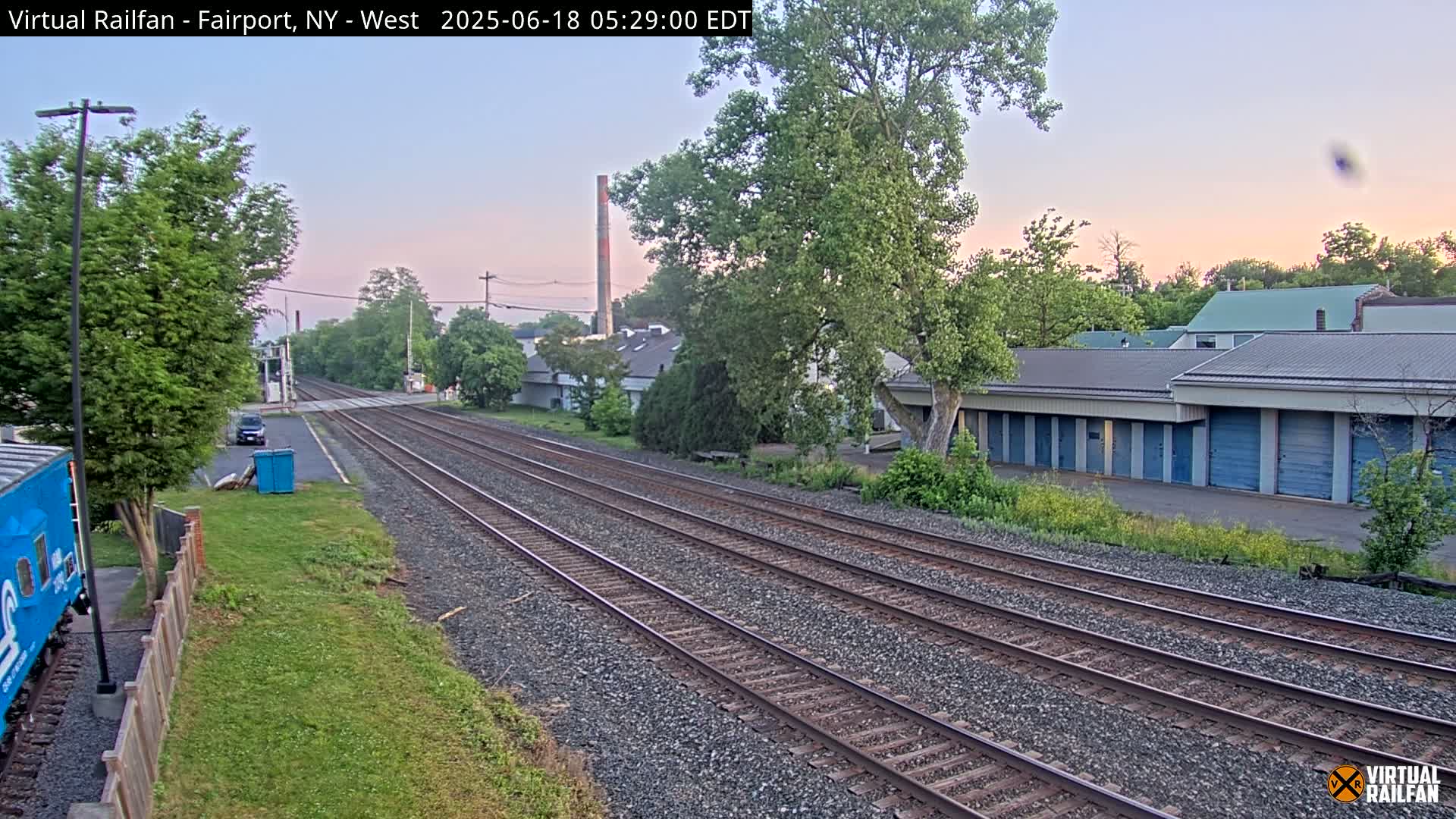 Three sets of train tracks extend into the distance under a pale sunrise, past a row of storage units and a small industrial building.