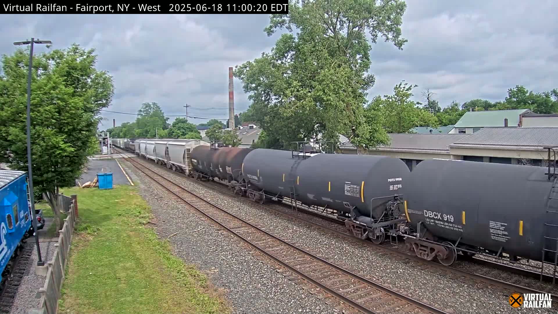 A long freight train, consisting of tanker cars and covered hopper cars, moves along a railroad track on an overcast day, passing by trees and buildings.