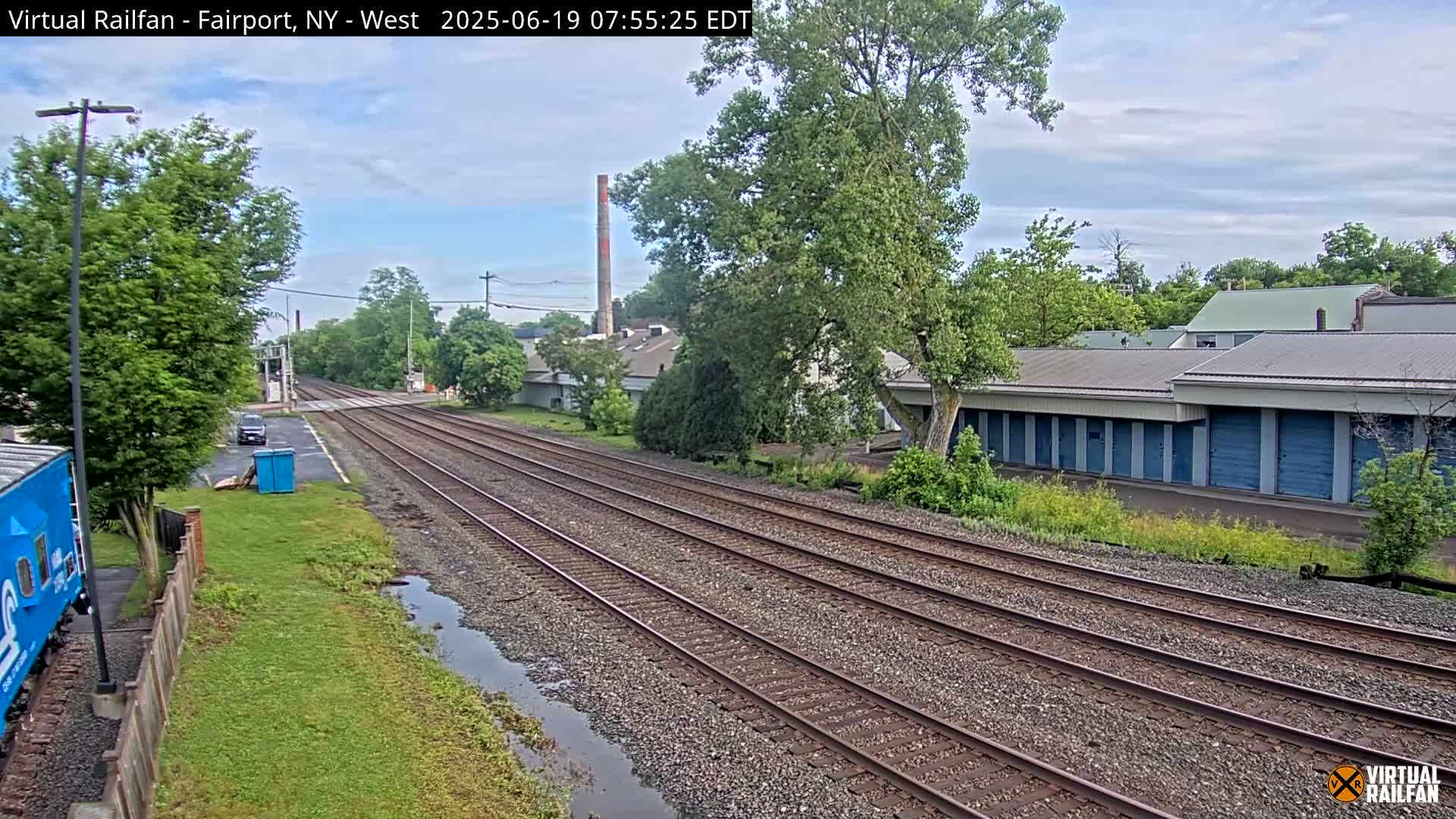 Multiple train tracks stretch into the distance on an overcast day, passing by industrial buildings and lush green vegetation alongside a road.