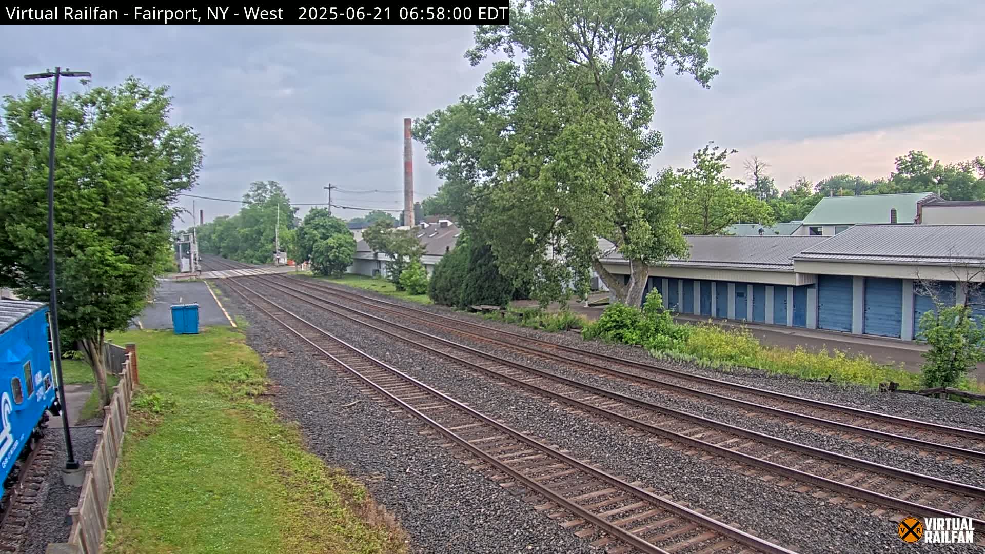Multiple train tracks extend into the distance under an overcast sky, past industrial buildings and lush green trees.