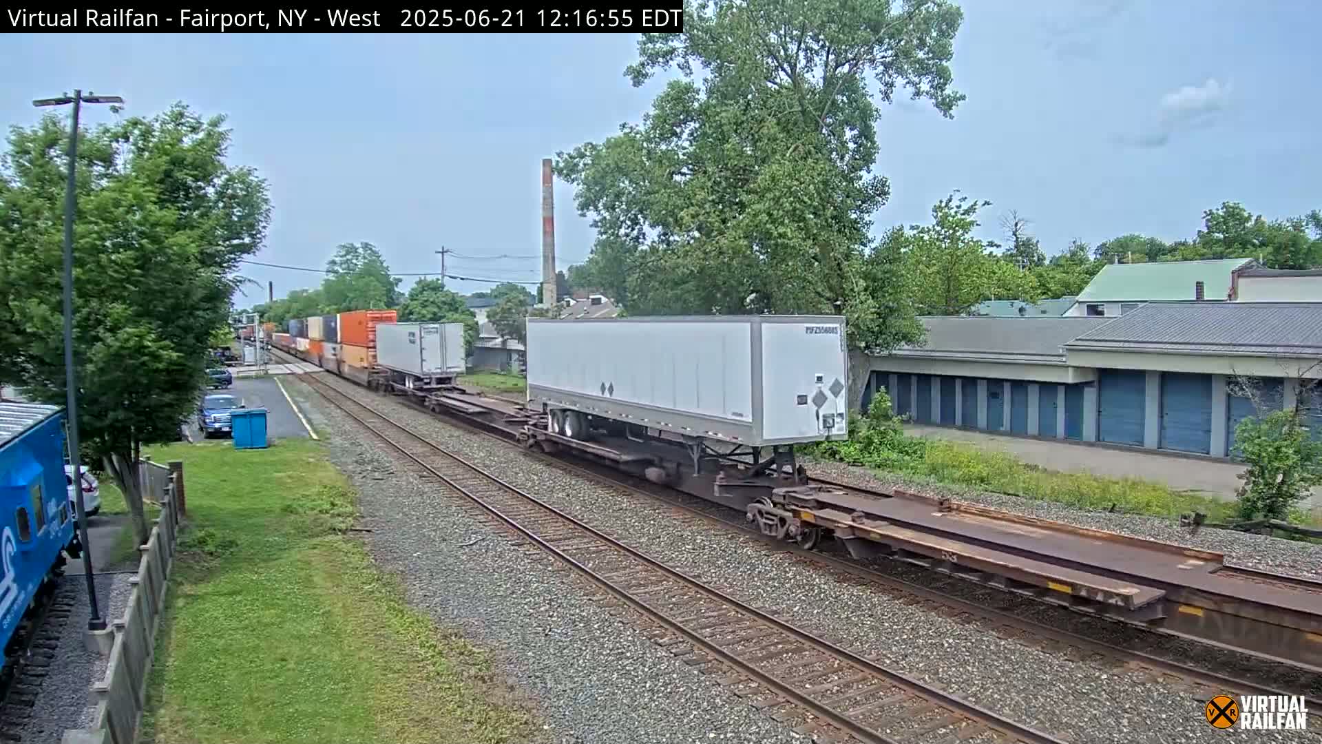 A freight train carrying shipping containers and a large white trailer moves along a railway track under a partly cloudy sky.