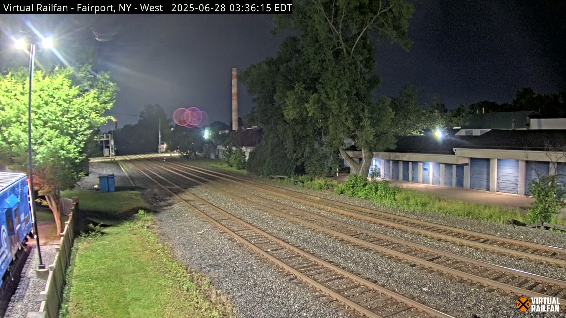 A nighttime view of train tracks extending into the distance, with a row of storage units and a tall smokestack visible in the background under a dark sky.