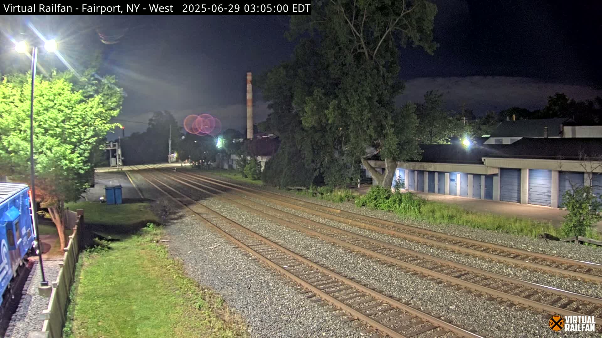 At night, under a cloudy sky, multiple train tracks extend into the distance, passing alongside a row of storage units and a tall smokestack.