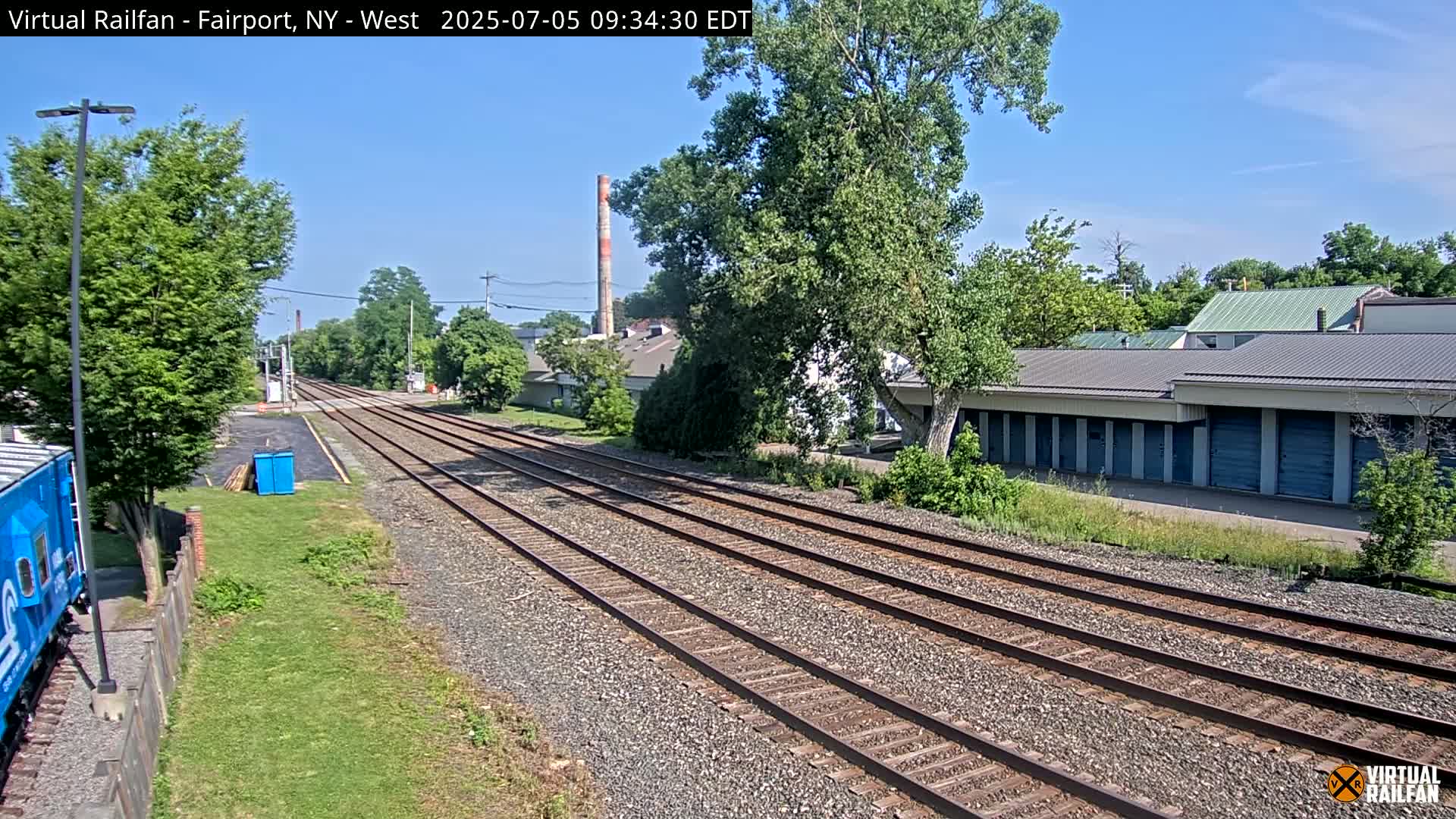 Multiple train tracks extend into the distance under a clear sky, past a row of storage units and some industrial buildings.