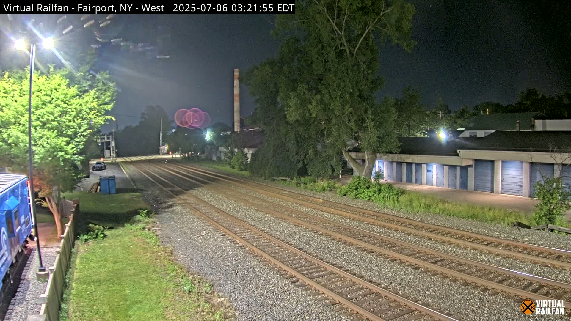At night, a train track with multiple sets of rails extends into the distance, past a row of storage units and under a mostly clear night sky.
