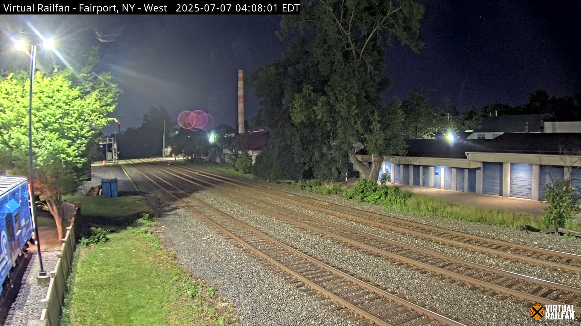 At night, multiple train tracks extend into the distance, passing alongside a row of storage units and a partially visible train car.