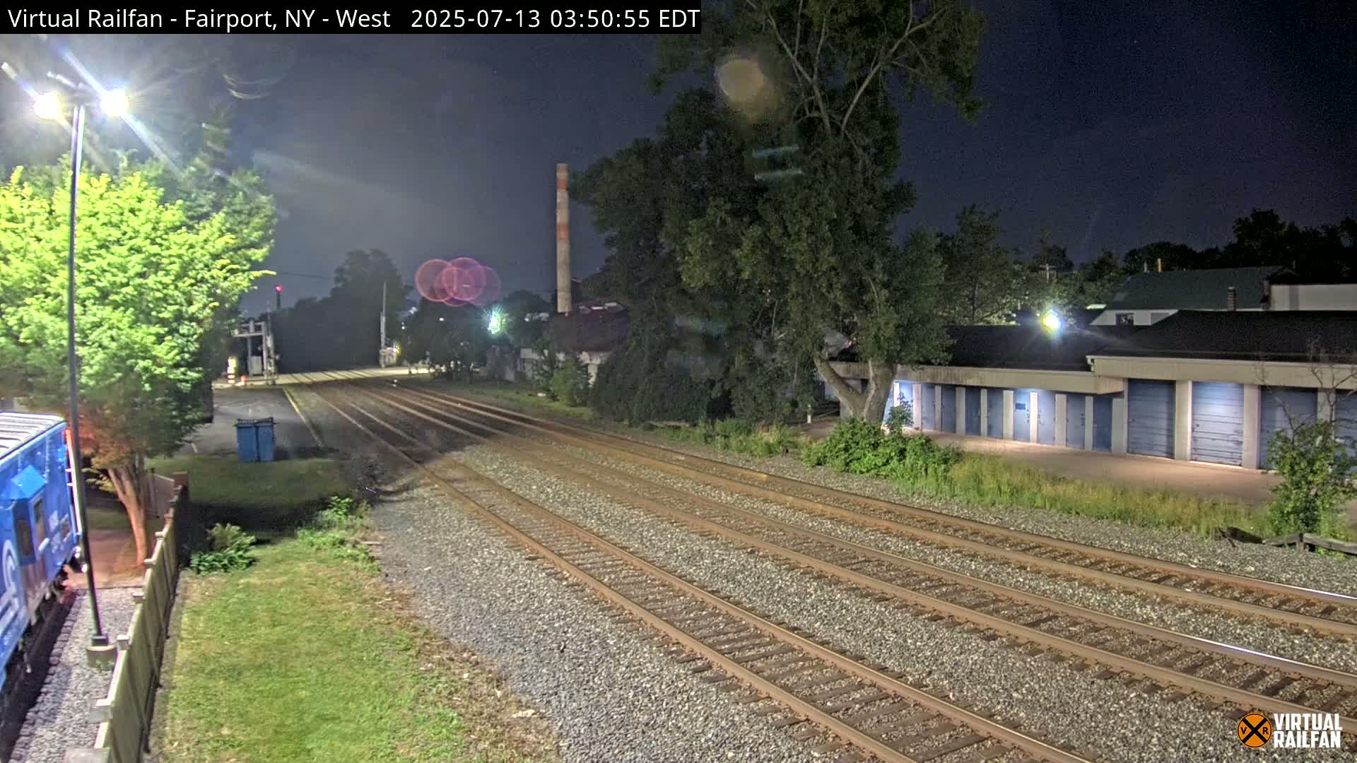 At night, a train and several parallel train tracks extend into the distance, passing alongside a row of storage units and trees under a dark sky.