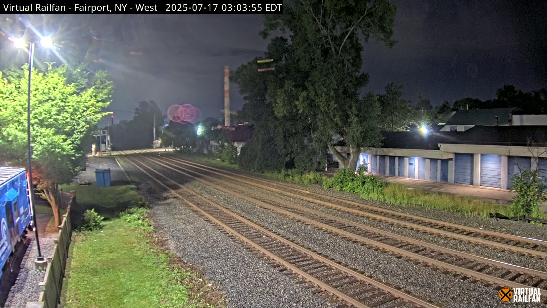 At night, multiple train tracks extend into the distance, passing beside a row of storage units and trees under a dark sky.