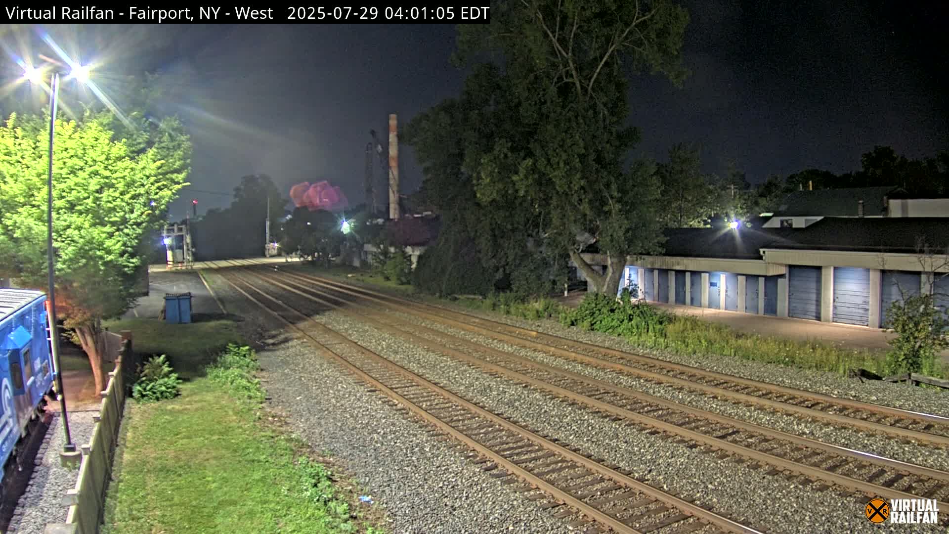 At night, multiple sets of train tracks extend into the distance, past a row of storage units and some trees, under a dark sky.