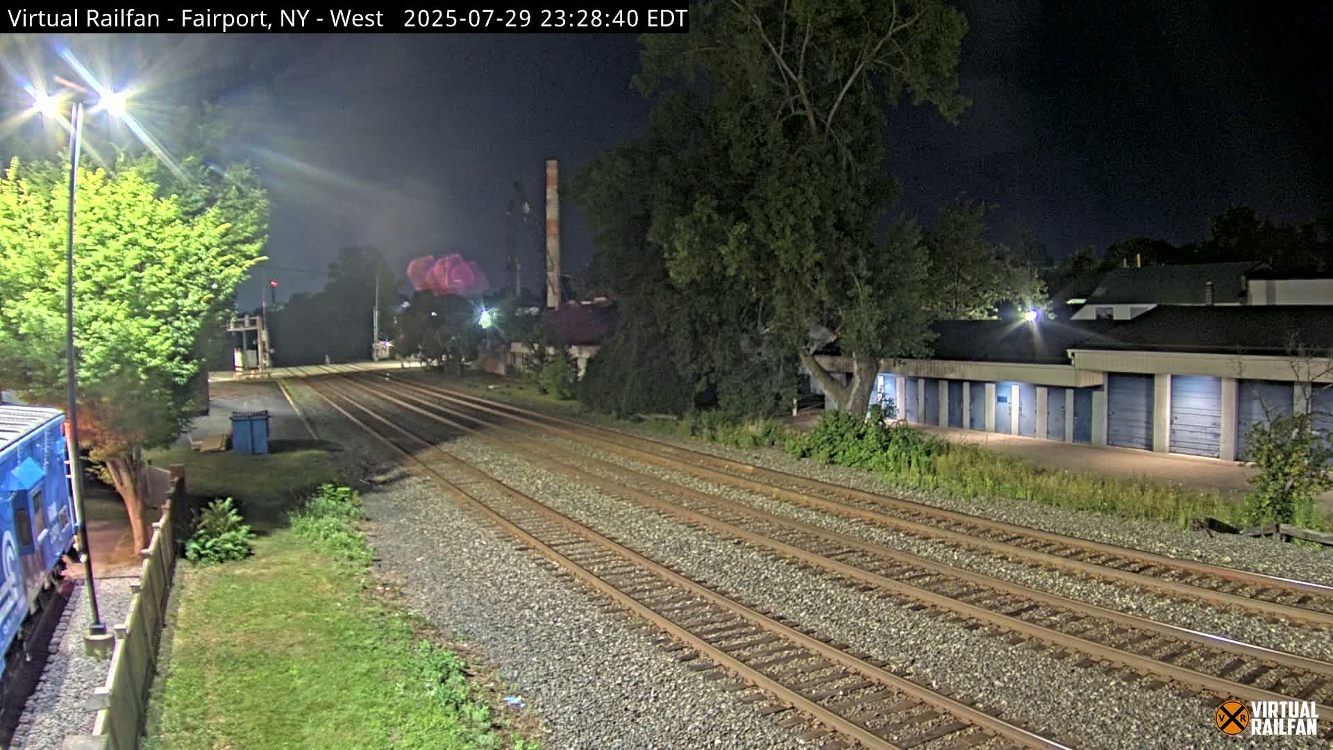 At night, multiple sets of train tracks extend into the distance, past a row of storage units and some trees, under a dark sky.