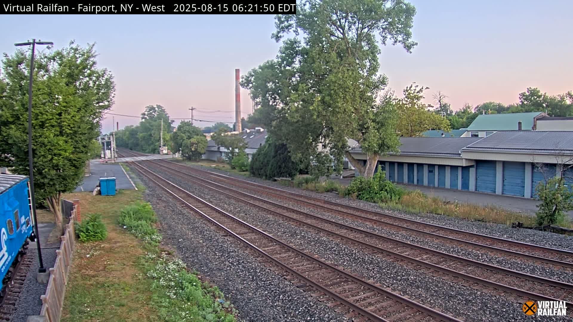 At night, multiple sets of train tracks extend into the distance, past a row of storage units and some trees, under a dark sky.