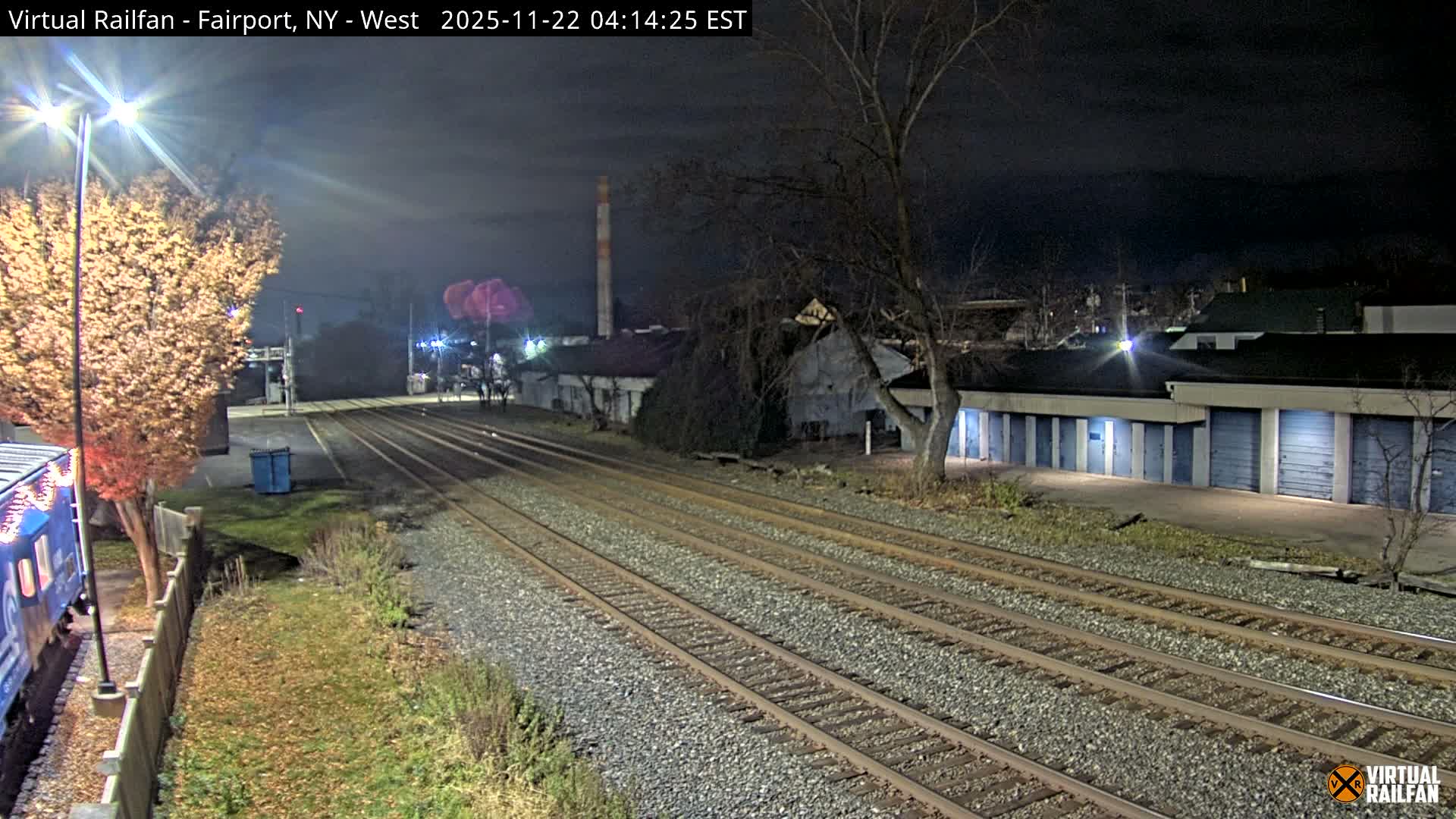 Multiple parallel railroad tracks stretch into the distance at night under a clear sky, bordered by a brightly lit tree and the front of a blue train car on the left, and a long building with rows of blue storage unit doors on the right, with an industrial chimney visible in the background.