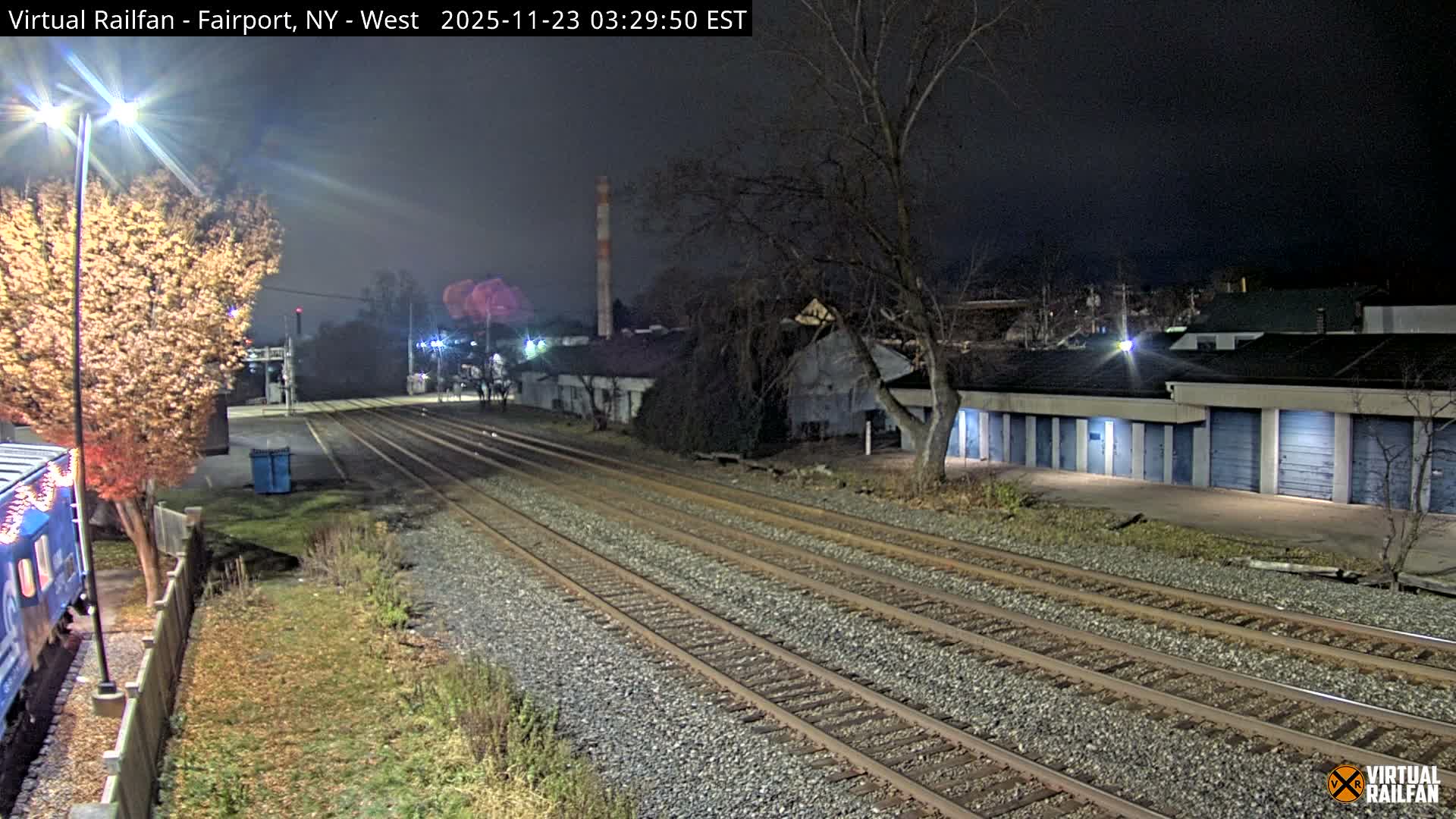 A nighttime view shows multiple railroad tracks stretching through an industrial area, bordered by a brightly lit autumn tree and a partial blue train car on the left, and a long building with numerous blue garage doors and a distant industrial chimney on the right, under a clear sky.