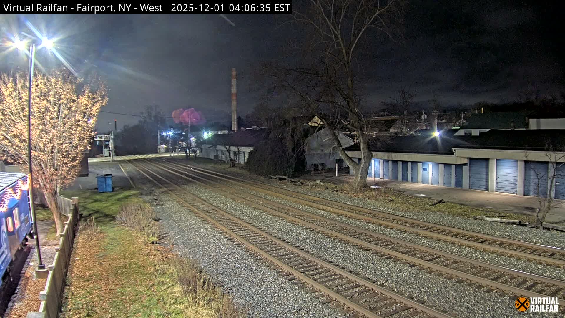 Multiple railroad tracks stretch into the distance at night, illuminated by artificial lights, with a partial view of a blue train car, bare trees, various buildings, and storage units under clear weather conditions.
