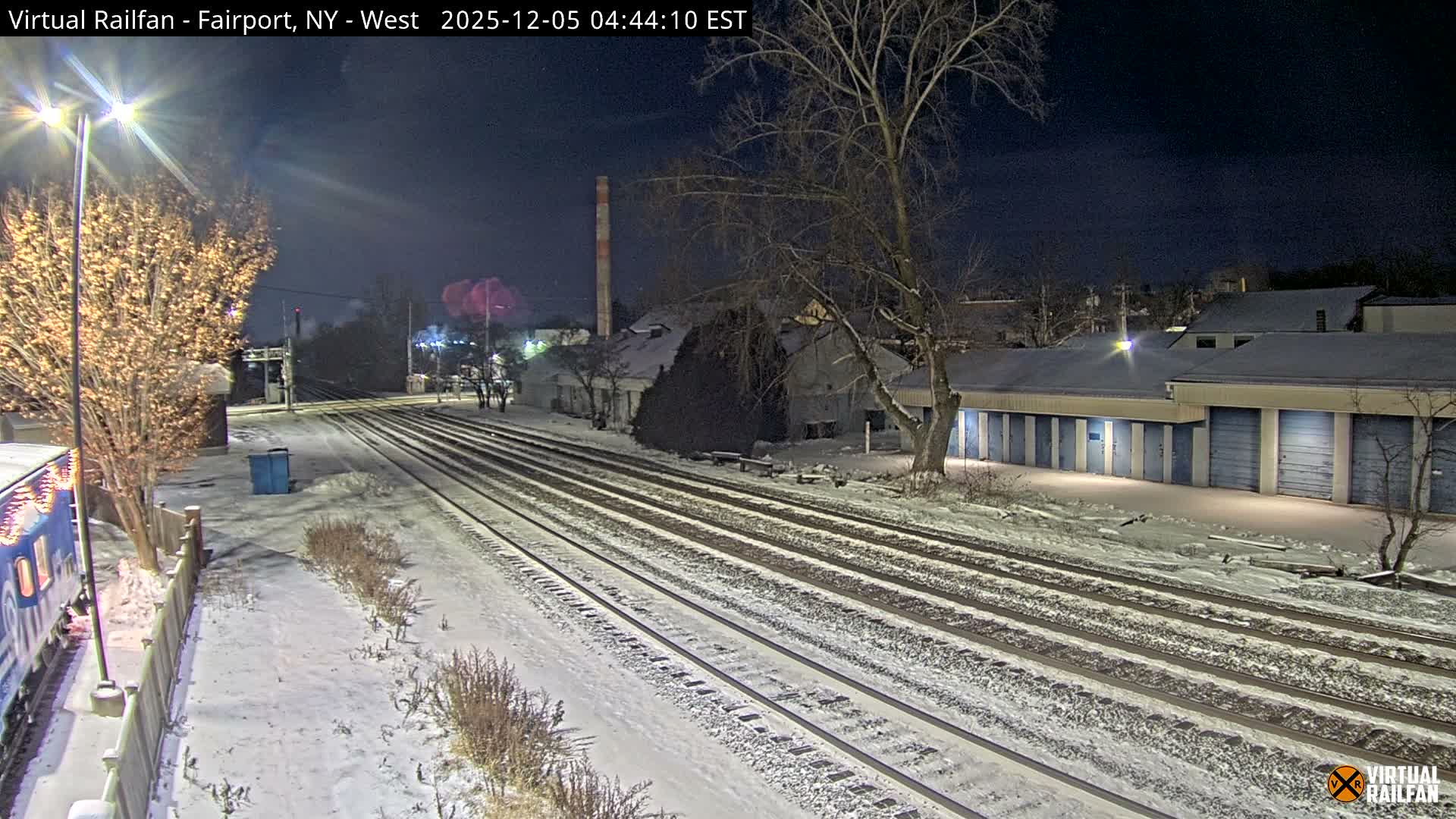 A snowy night scene depicts multiple parallel train tracks, bare trees, and buildings, with a train car visible on the left, all illuminated by streetlights under a clear, dark sky.