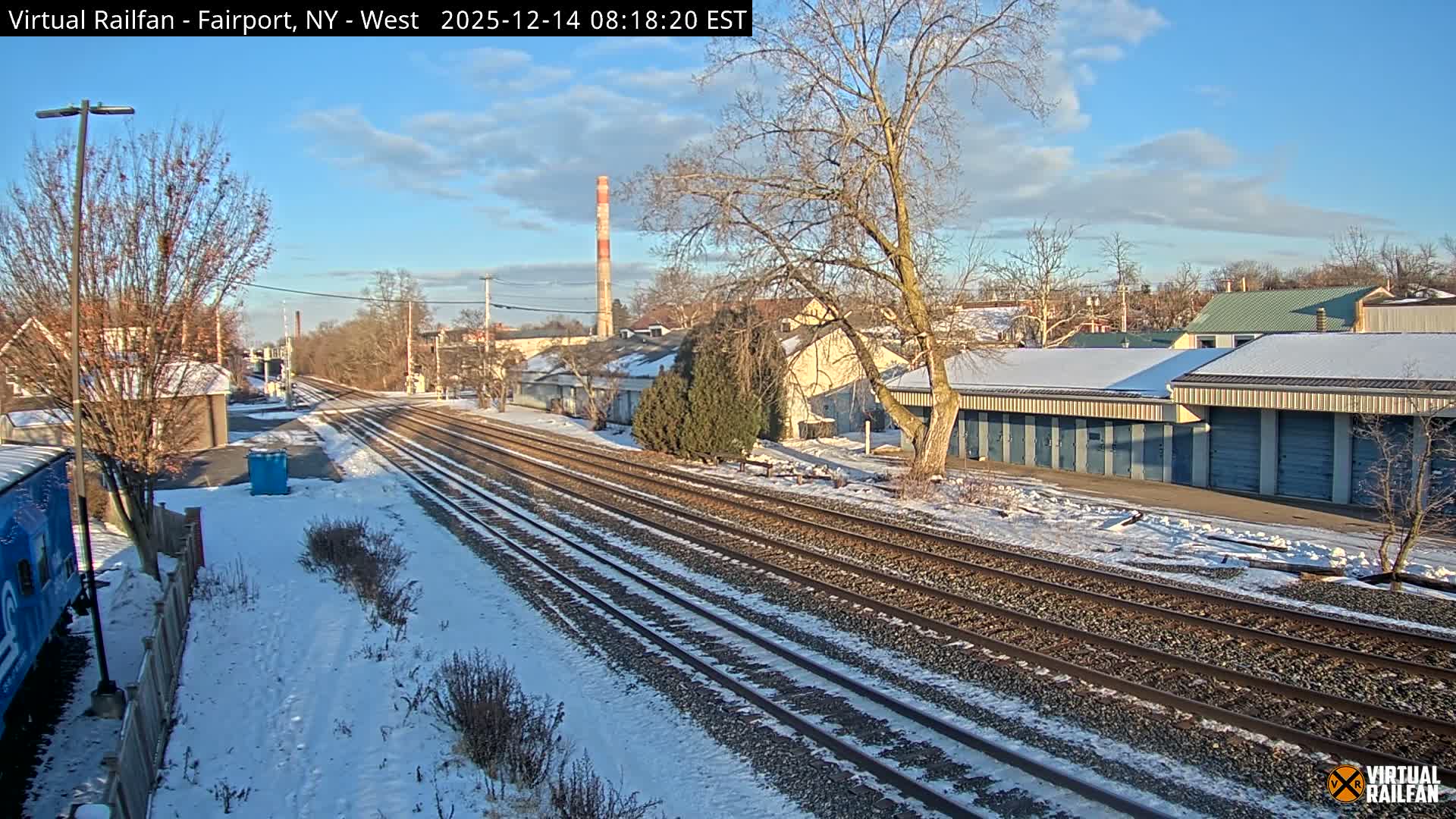 A snowy night scene depicts multiple parallel train tracks, bare trees, and buildings, with a train car visible on the left, all illuminated by streetlights under a clear, dark sky.