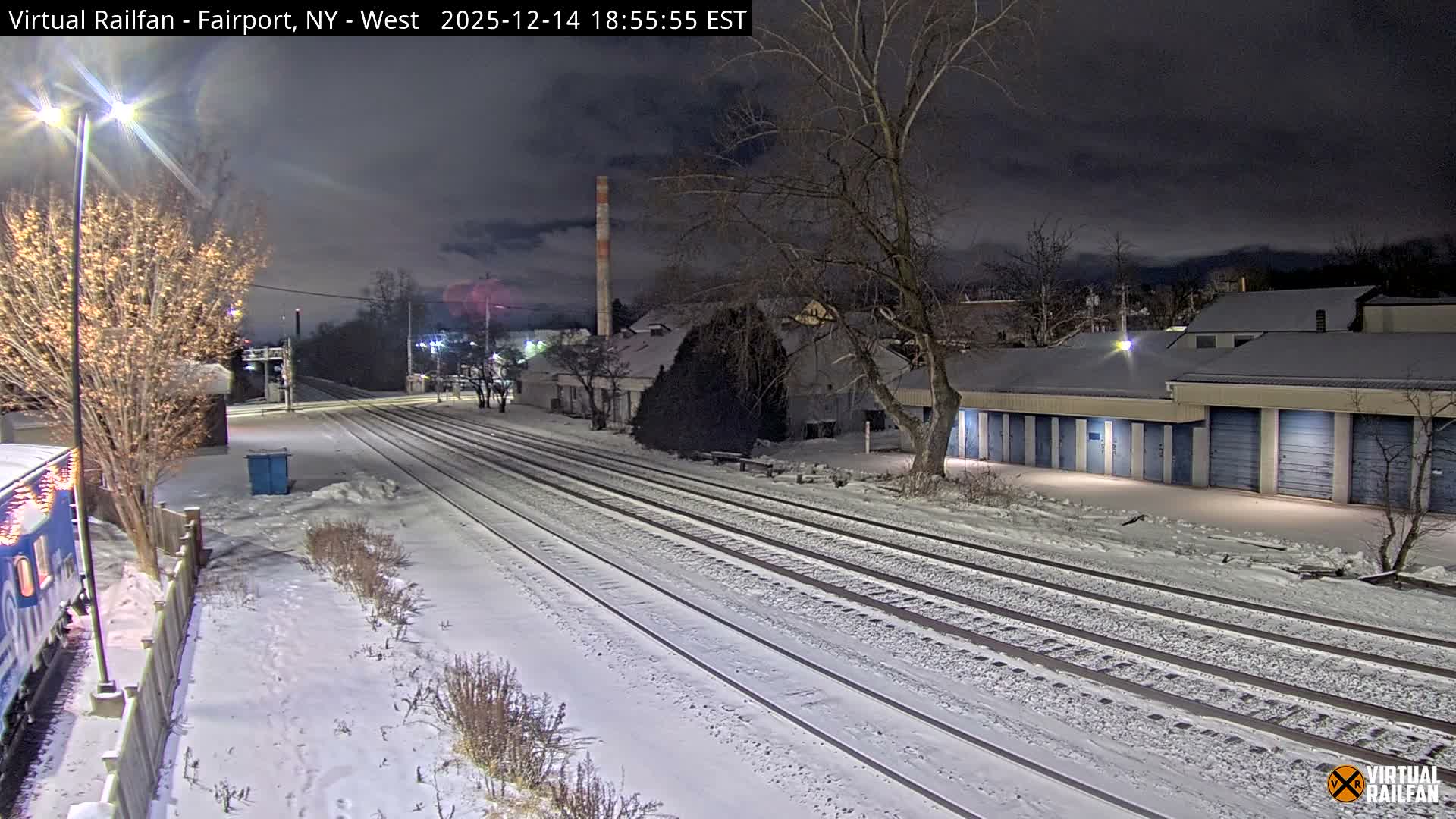 A snowy night scene depicts multiple parallel train tracks, bare trees, and buildings, with a train car visible on the left, all illuminated by streetlights under a clear, dark sky.