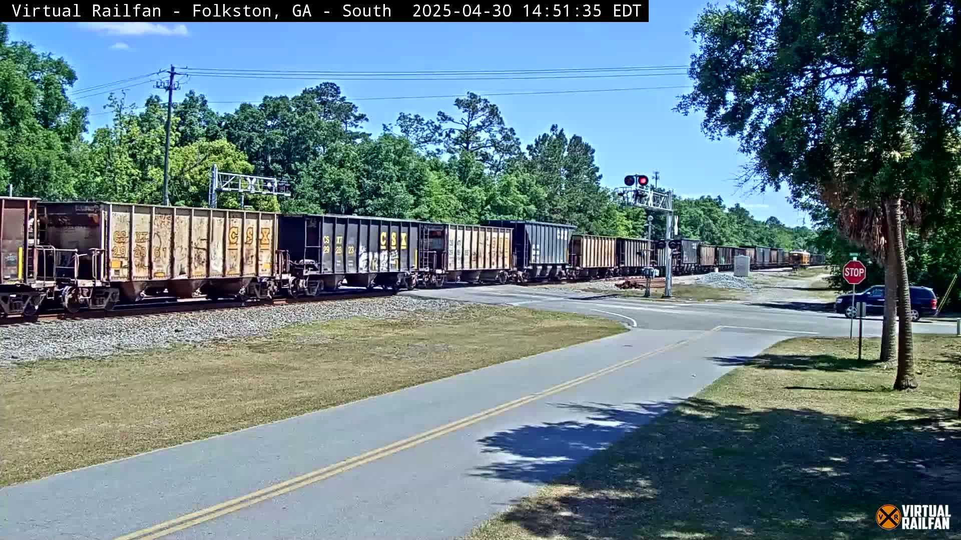 A long freight train passes a road crossing under a sunny sky, with green trees and grass surrounding the area.