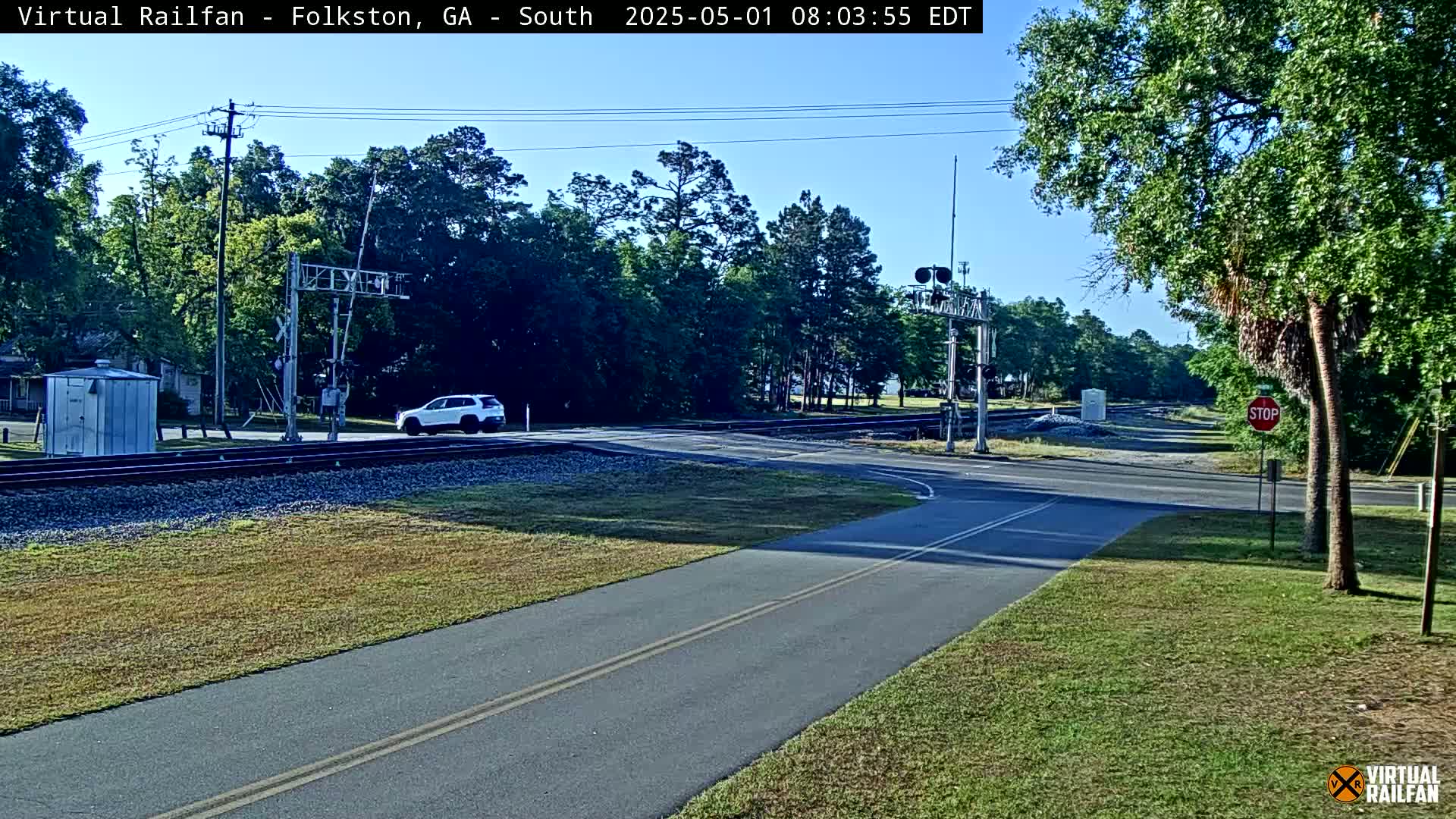 A paved road intersects with a railroad crossing under a sunny sky, with a white SUV visible near the crossing and lush green vegetation surrounding the area.