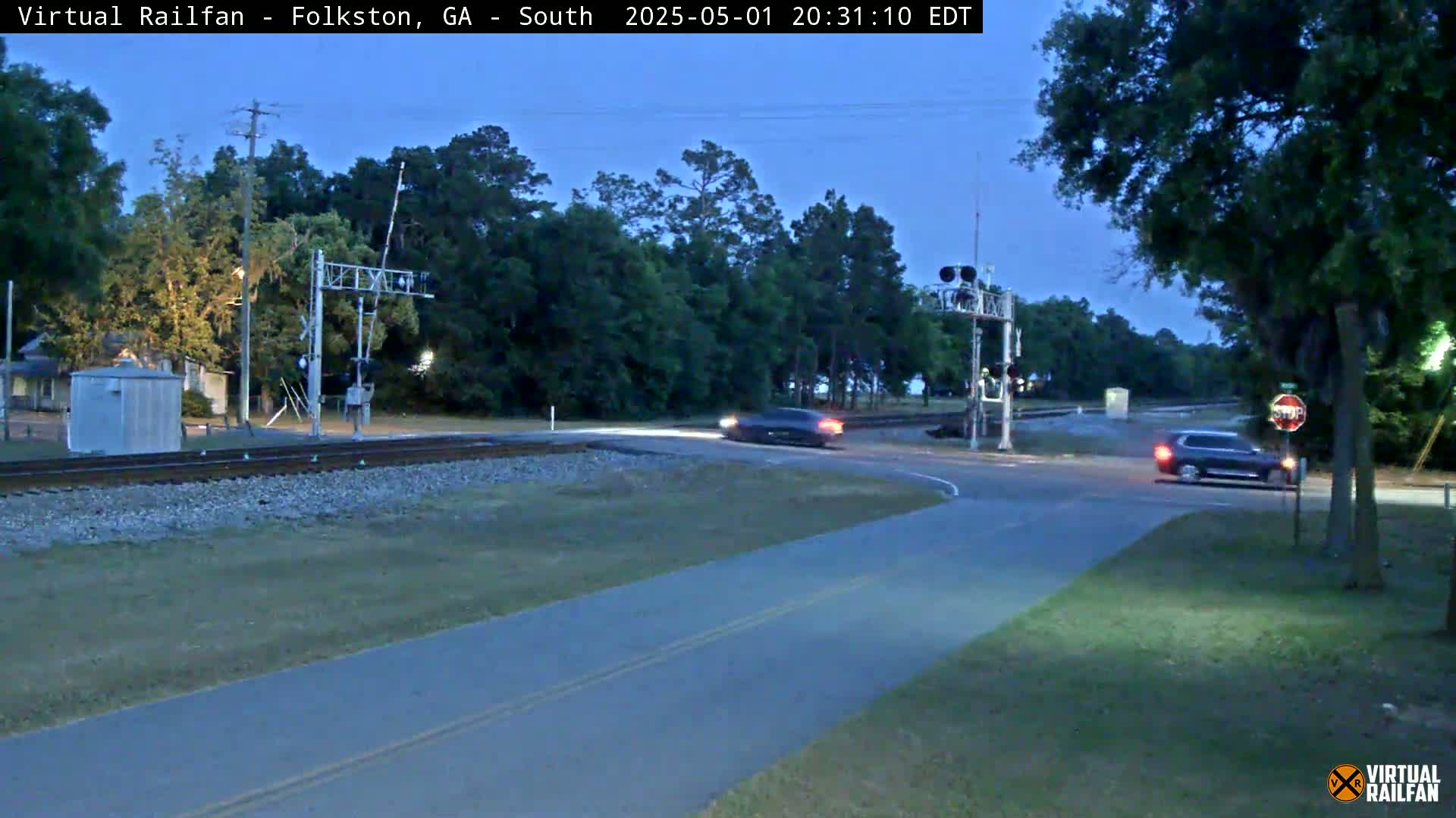 A road and railroad crossing intersect under a twilight sky, with two vehicles visible near the intersection.