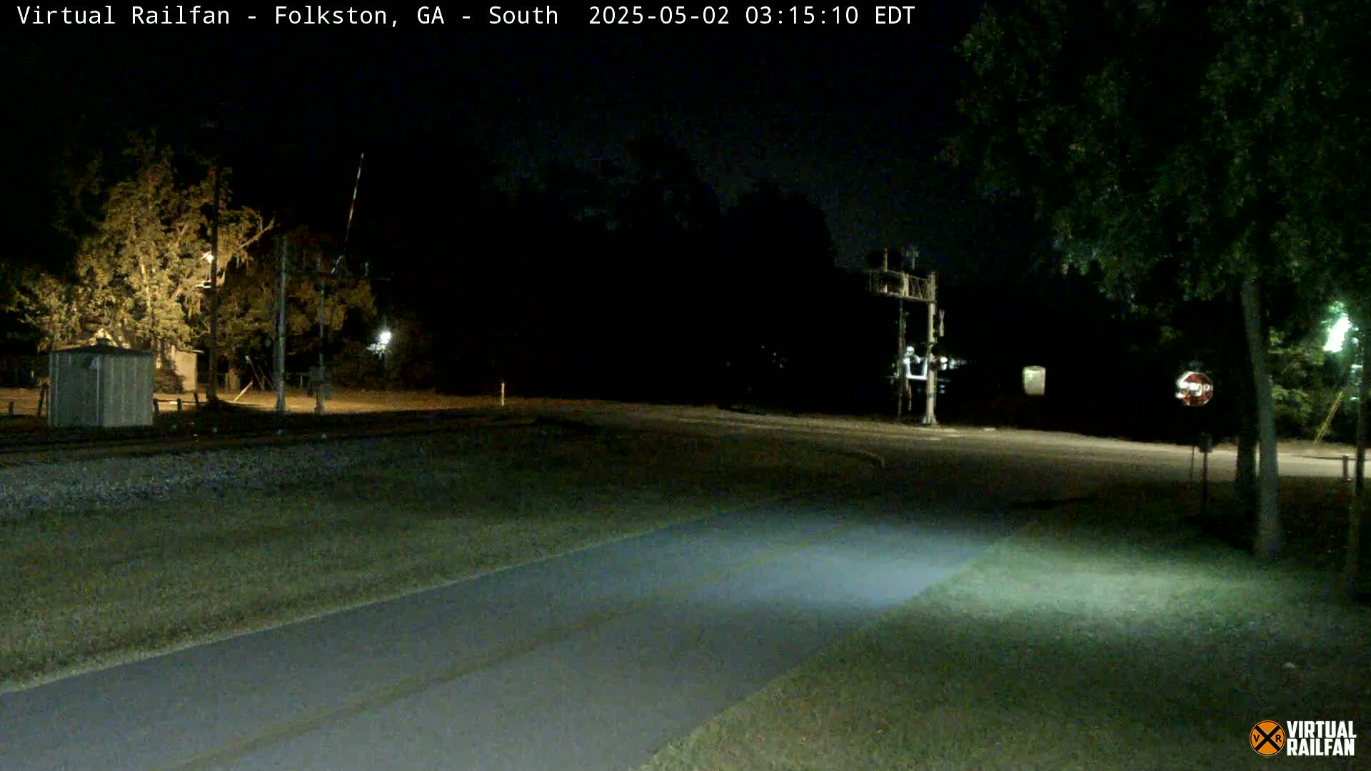 A nighttime view of a railroad crossing with a paved path leading toward it, surrounded by trees and under artificial light.