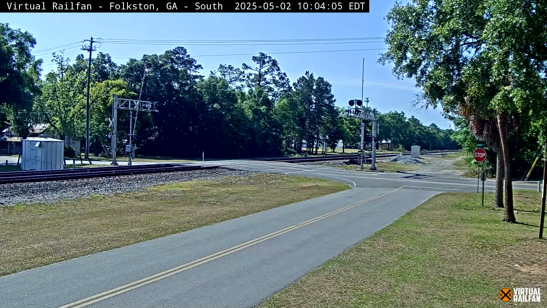 A paved road intersects with a railroad crossing under a sunny sky, surrounded by green vegetation and trees.