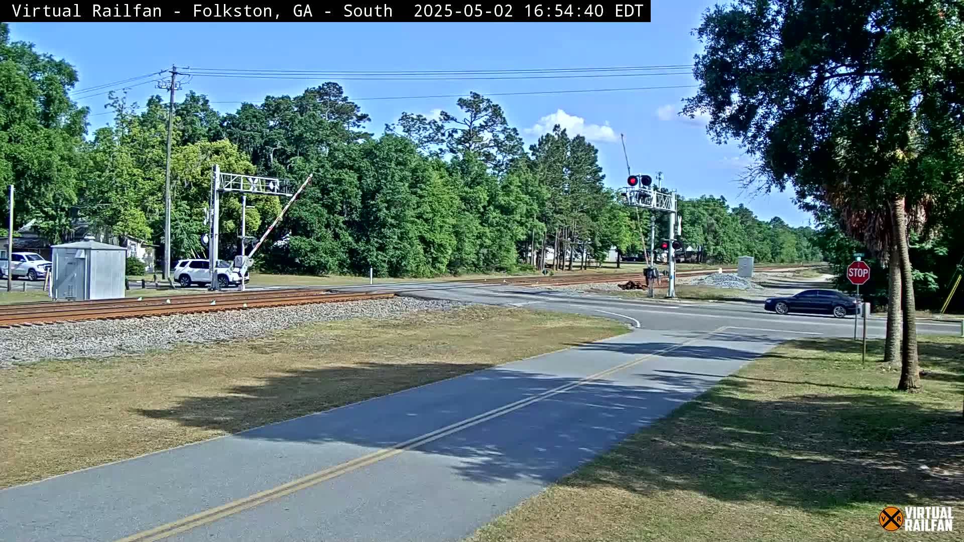 A road intersects a railroad track under a mostly sunny sky, with cars present on both the road and a nearby parking area, surrounded by green trees and vegetation.