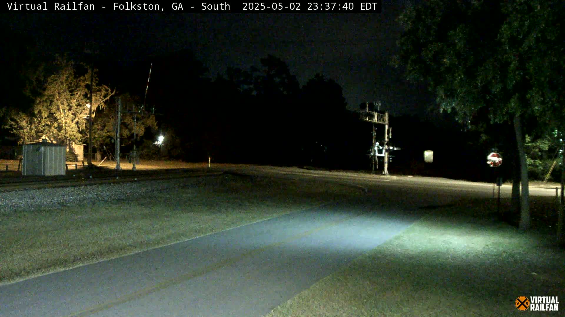 A nighttime view of a railroad crossing with a paved path, trees, and a small building under dark, clear skies.