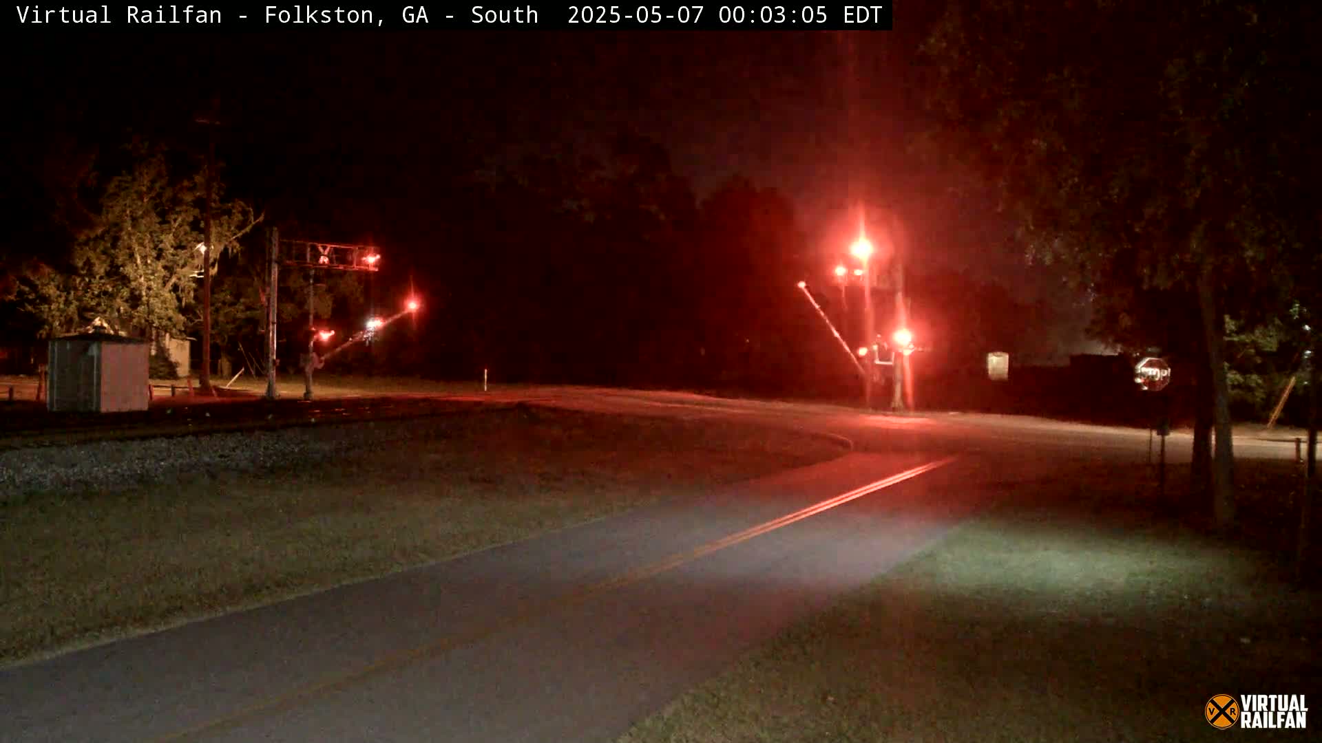 A nighttime view of a railroad crossing with flashing red lights, a paved path, and trees surrounding the area.