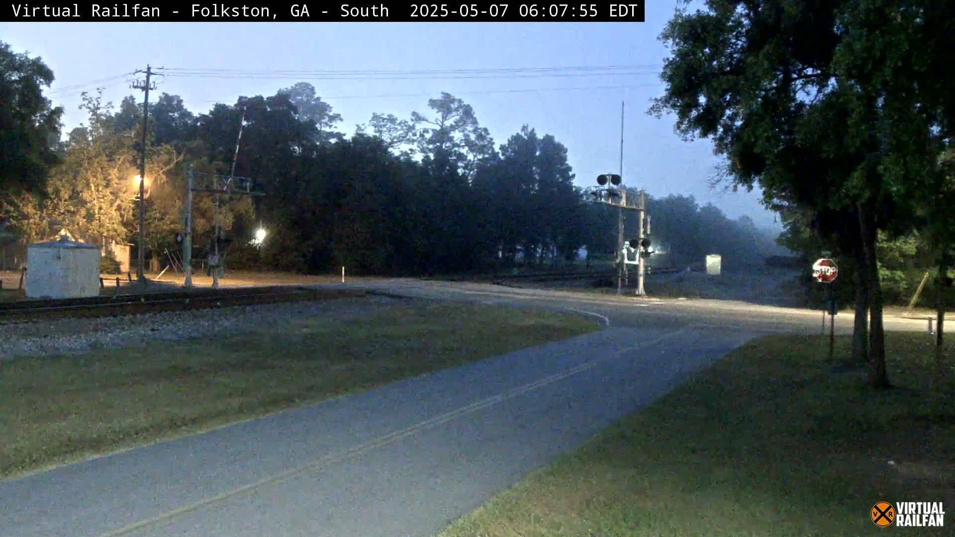 A road intersects with a railroad crossing at twilight under foggy conditions, with trees lining the edges of the road and railroad tracks.